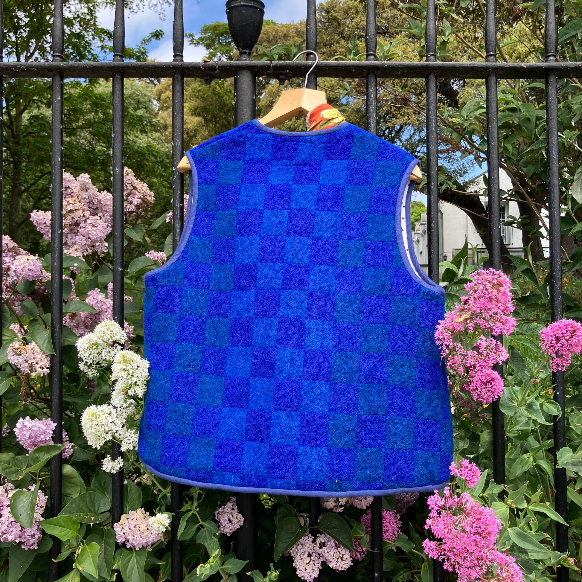 Vest/gilet/waistcoat made from a reclaimed blue checkerboard weave Irish wool Gaiety blanket, photographed in front of a lilac bush (back view)