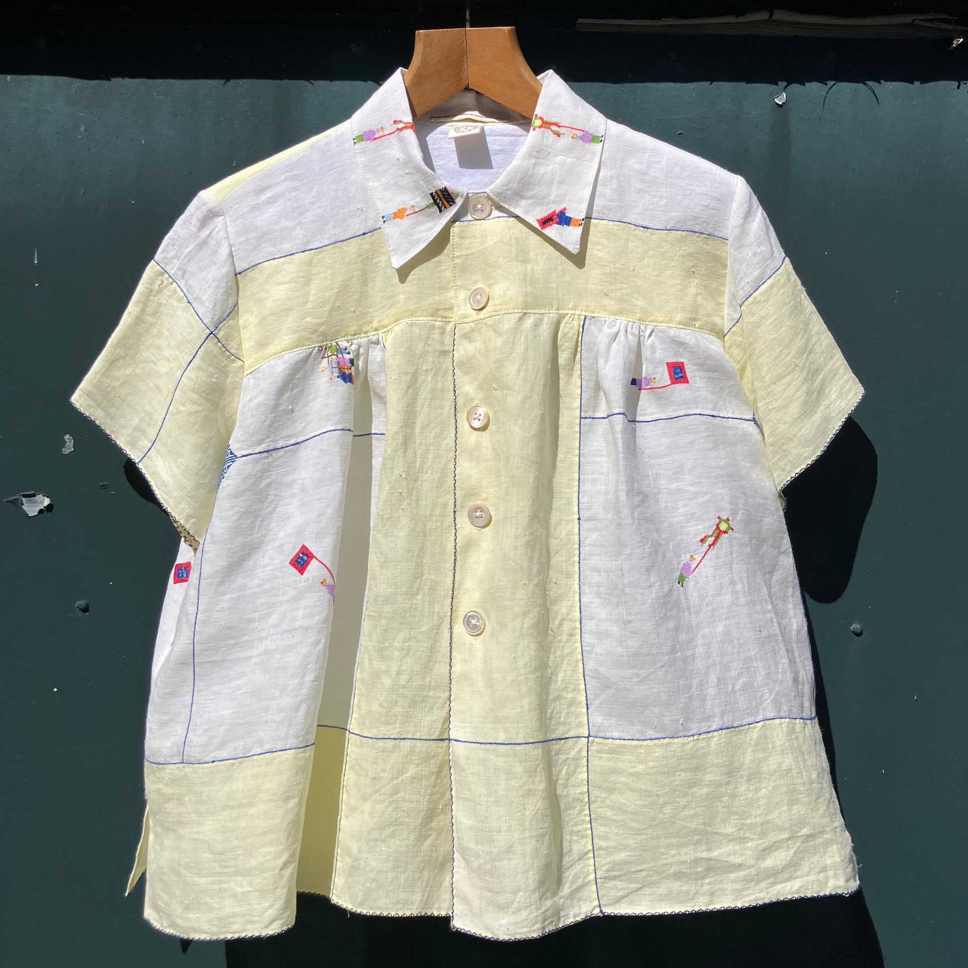 Shirt made from a found vintage tablecloth with a parade of small embroidered figures holding signs, kites and lanterns