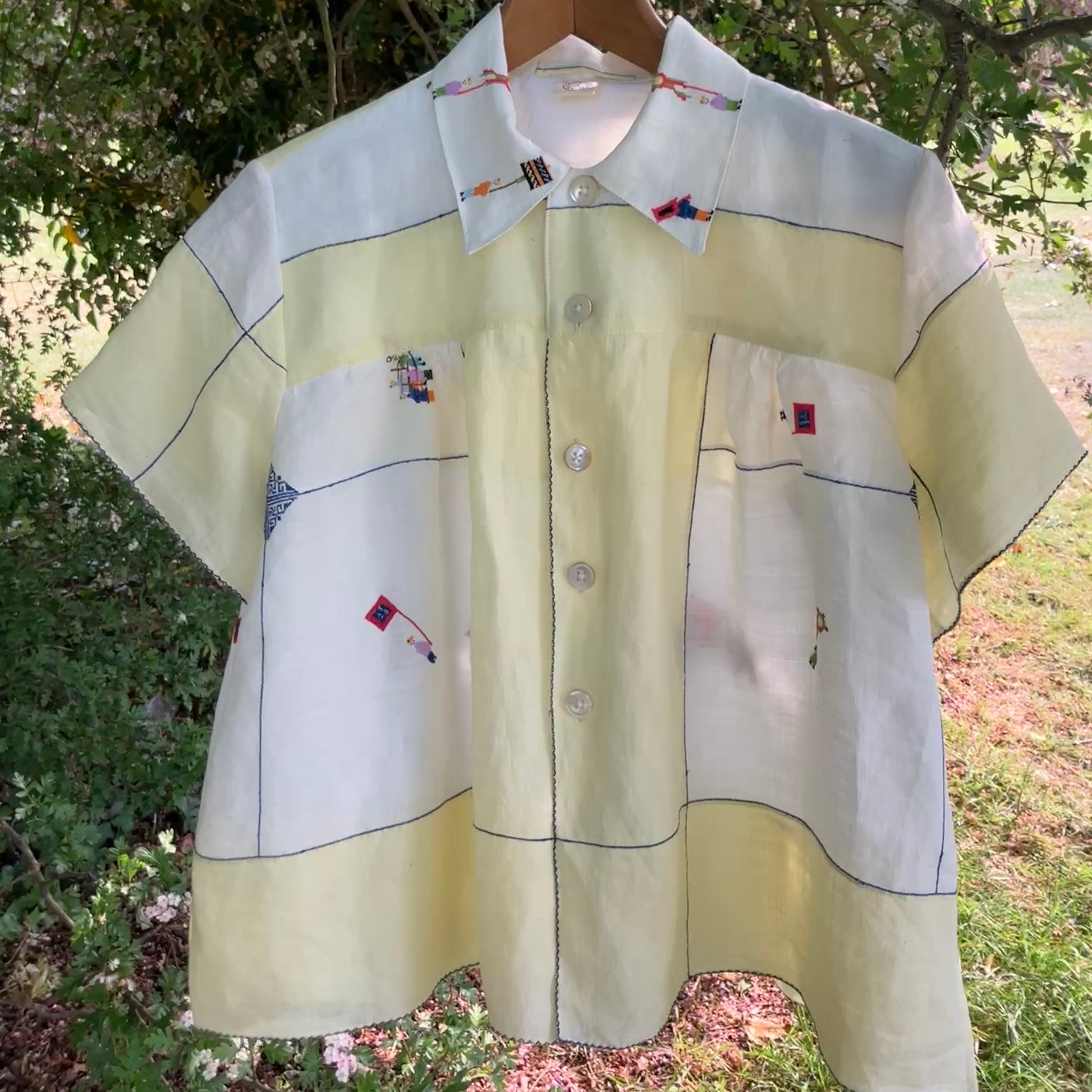 Shirt made from a found vintage tablecloth with a parade of small embroidered figures holding signs, kites and lanterns