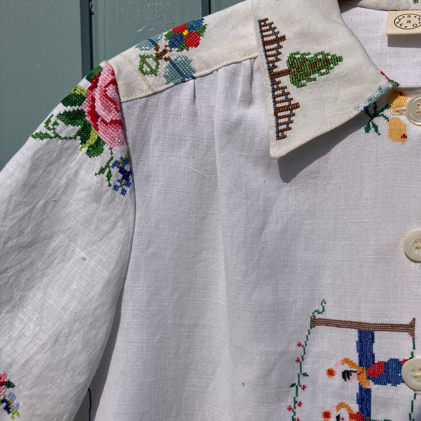 Linen shirt made from a patchwork of recycled linen tablecloths with cross-stitch motifs including dancers, flowers, trees, lemons, tomatoes and people in a horse-drawn chariot