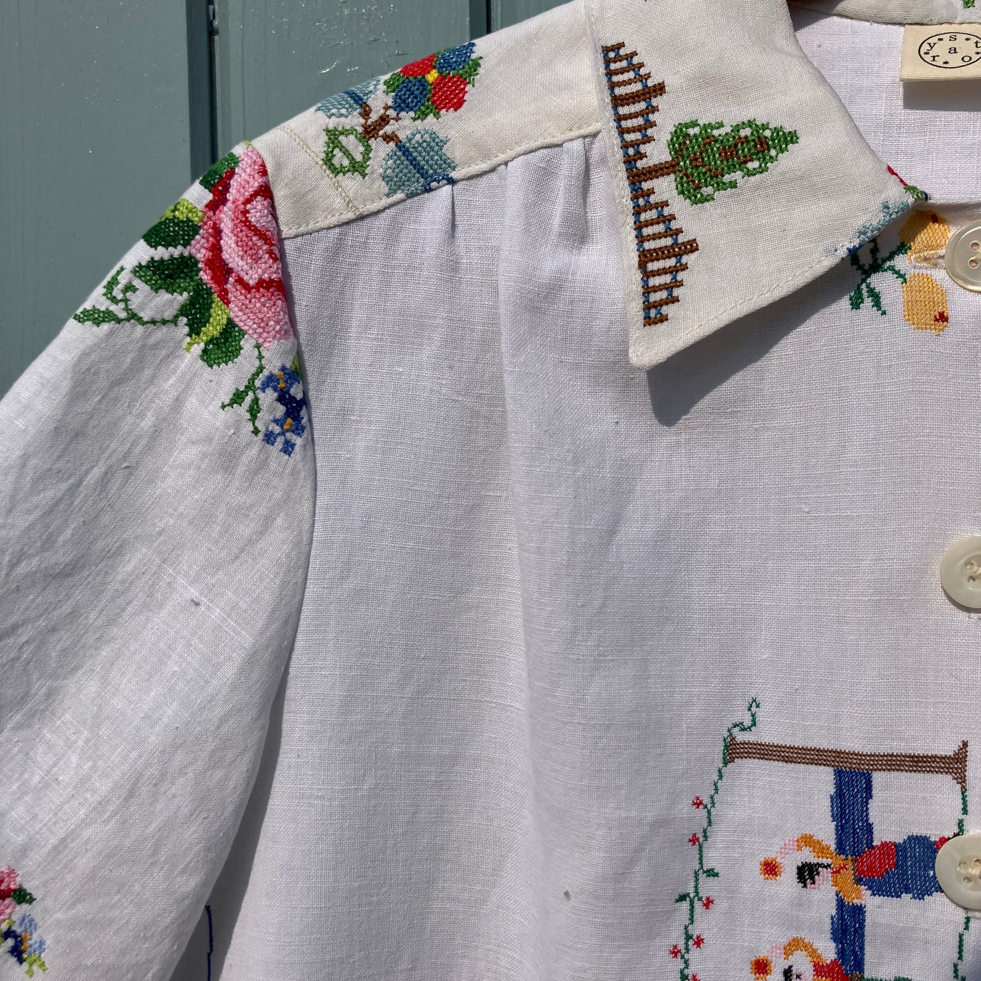 Linen shirt made from a patchwork of recycled linen tablecloths with cross-stitch motifs including dancers, flowers, trees, lemons, tomatoes and people in a horse-drawn chariot