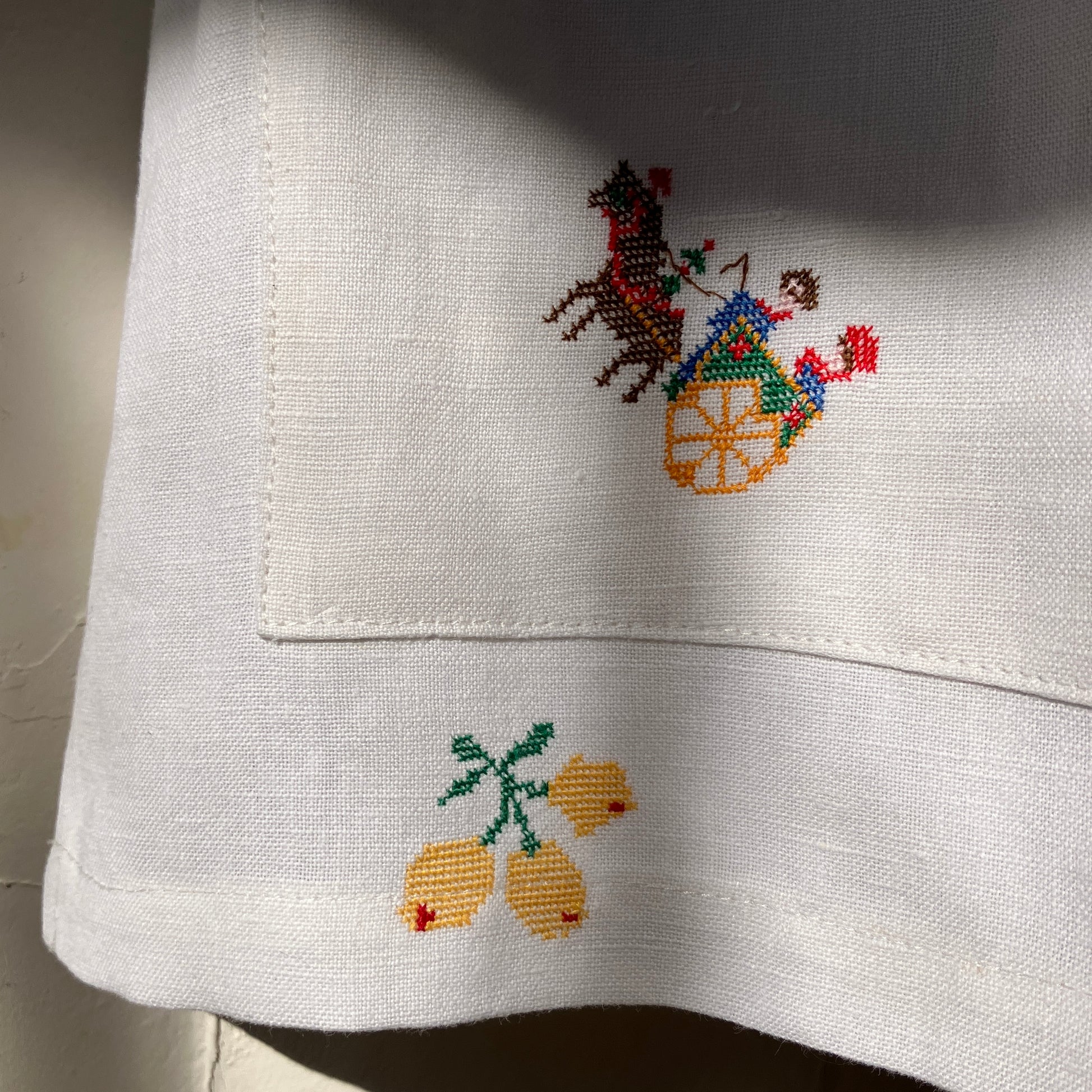 Linen shirt made from a patchwork of recycled linen tablecloths with cross-stitch motifs including dancers, flowers, trees, lemons, tomatoes and people in a horse-drawn chariot