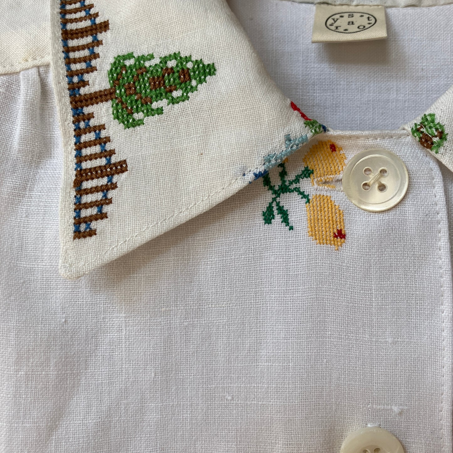 Linen shirt made from a patchwork of recycled linen tablecloths with cross-stitch motifs including dancers, flowers, trees, lemons, tomatoes and people in a horse-drawn chariot