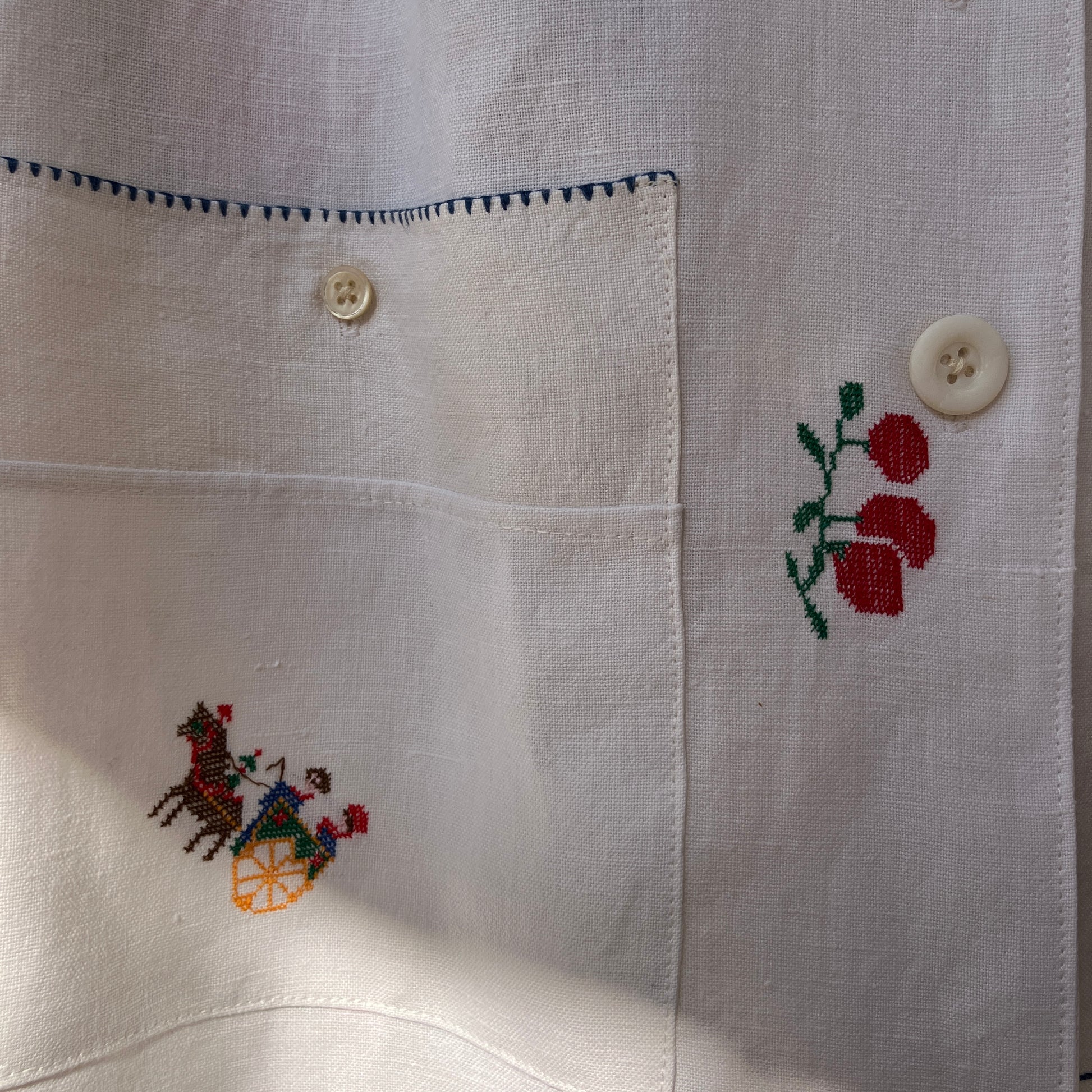 Linen shirt made from a patchwork of recycled linen tablecloths with cross-stitch motifs including dancers, flowers, trees, lemons, tomatoes and people in a horse-drawn chariot