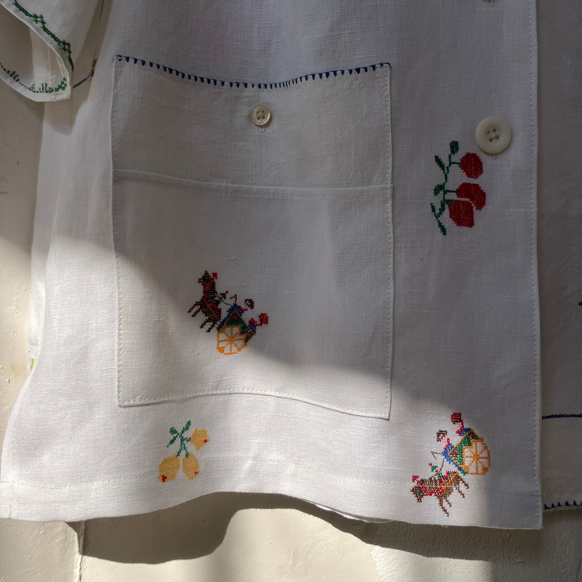 Linen shirt made from a patchwork of recycled linen tablecloths with cross-stitch motifs including dancers, flowers, trees, lemons, tomatoes and people in a horse-drawn chariot