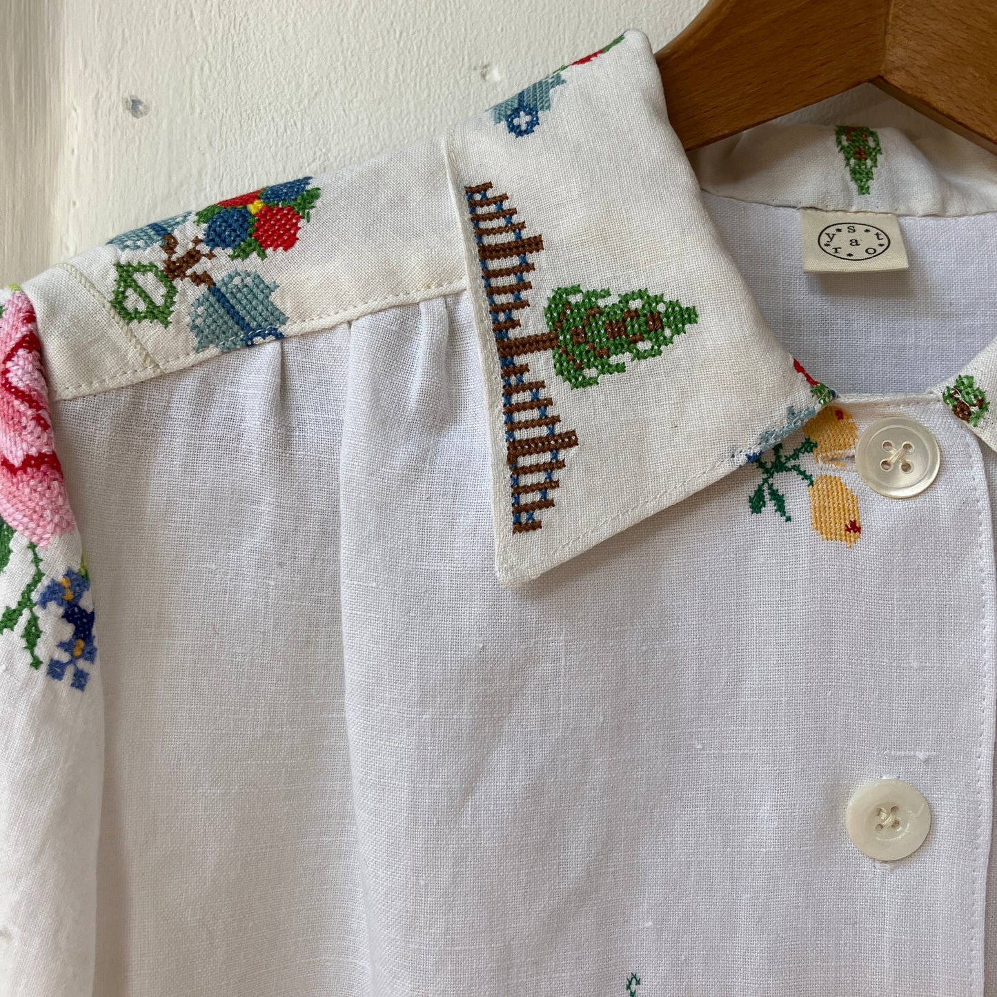 Linen shirt made from a patchwork of recycled linen tablecloths with cross-stitch motifs including dancers, flowers, trees, lemons, tomatoes and people in a horse-drawn chariot