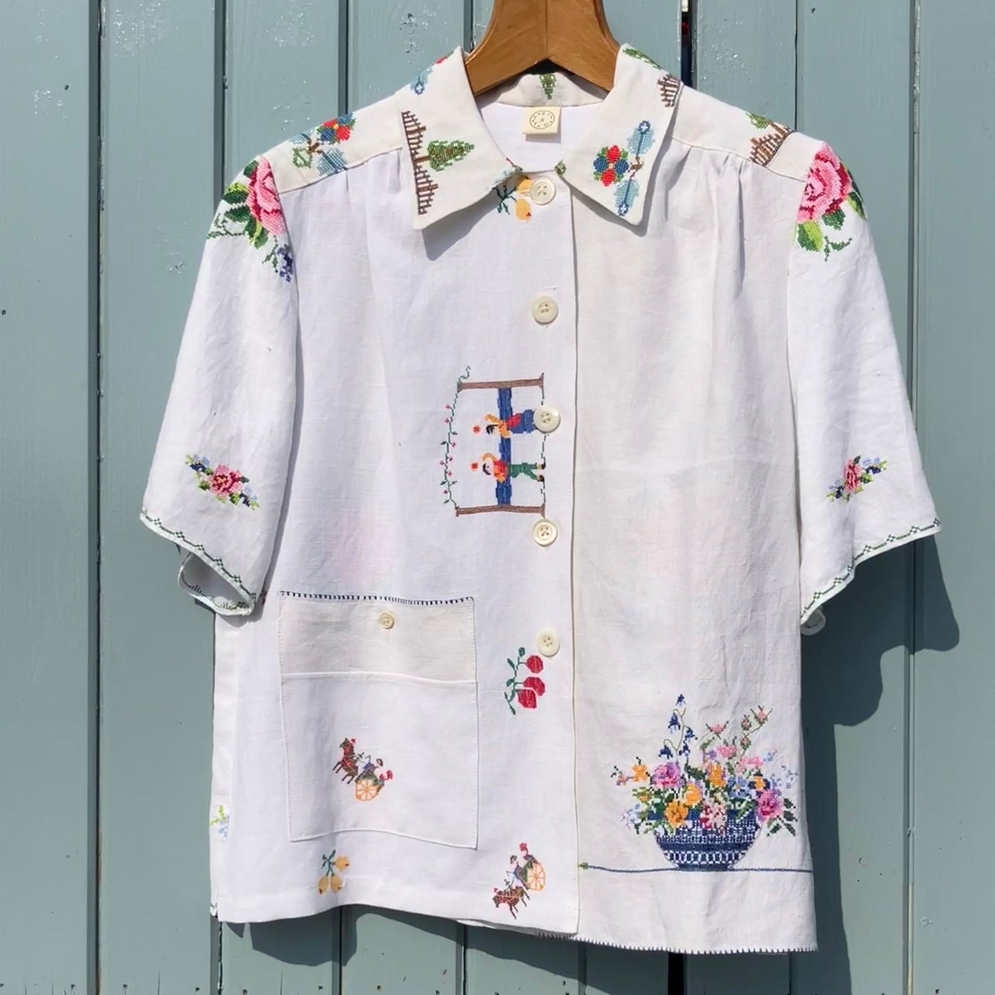 Linen shirt made from a patchwork of recycled linen tablecloths with cross-stitch motifs including dancers, flowers, trees, lemons, tomatoes and people in a horse-drawn chariot