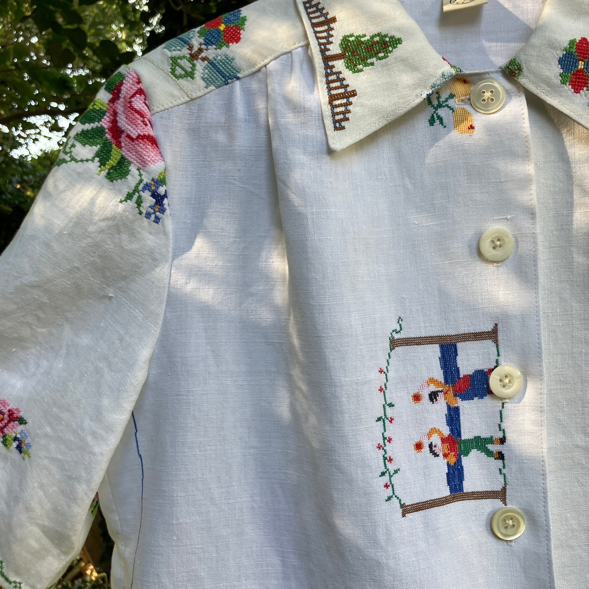 Linen shirt made from a patchwork of recycled linen tablecloths with cross-stitch motifs including dancers, flowers, trees, lemons, tomatoes and people in a horse-drawn chariot