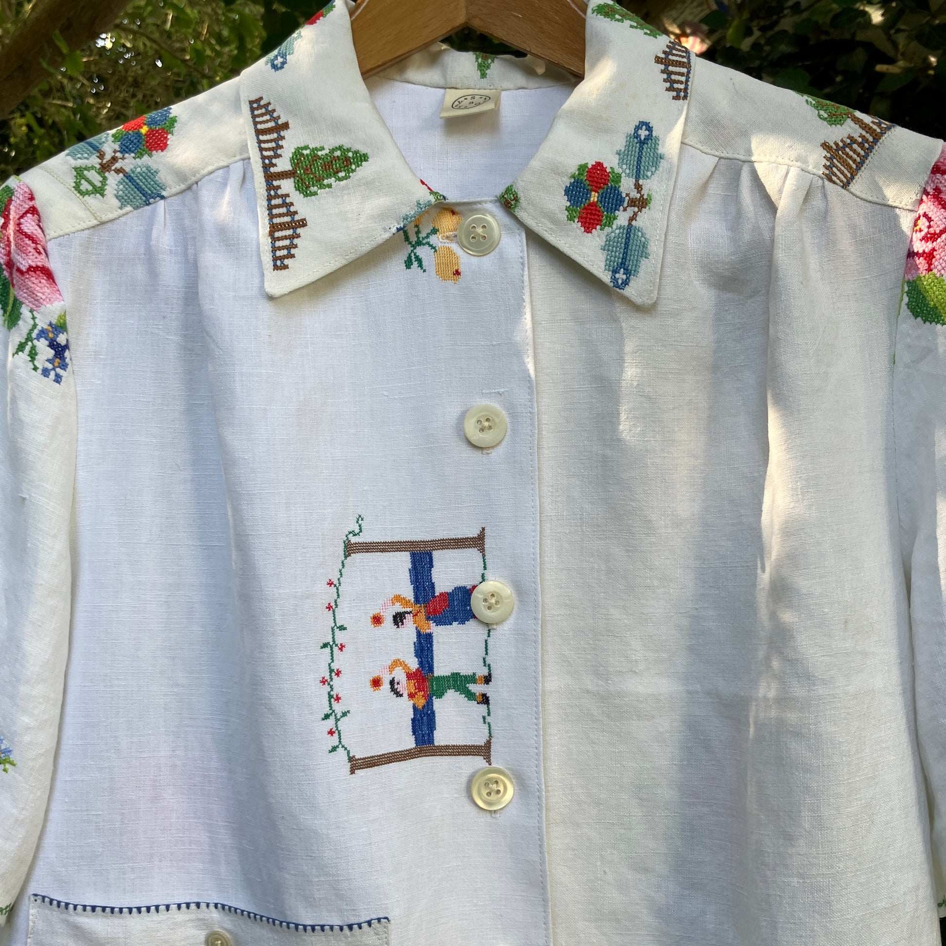 Linen shirt made from a patchwork of recycled linen tablecloths with cross-stitch motifs including dancers, flowers, trees, lemons, tomatoes and people in a horse-drawn chariot