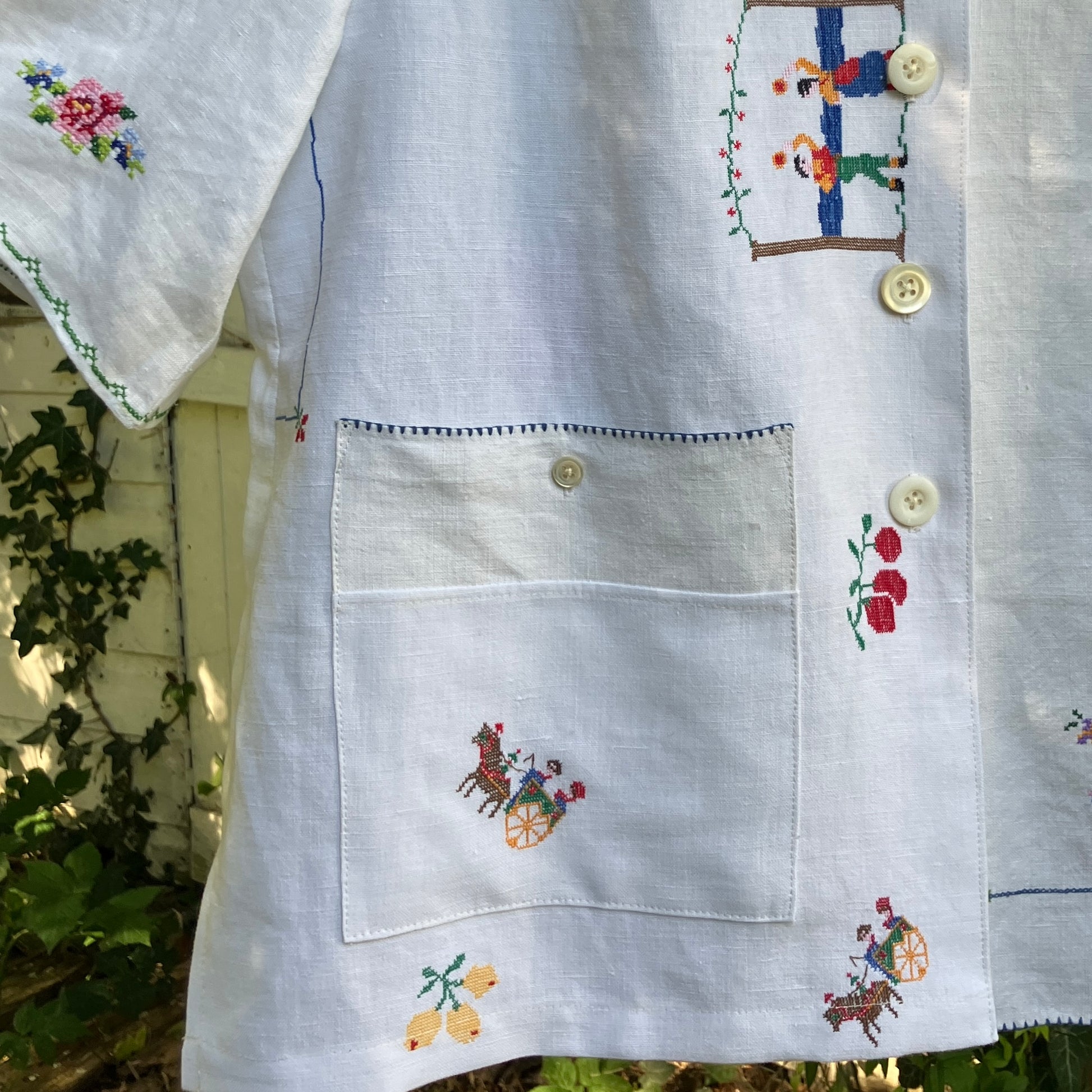 Linen shirt made from a patchwork of recycled linen tablecloths with cross-stitch motifs including dancers, flowers, trees, lemons, tomatoes and people in a horse-drawn chariot