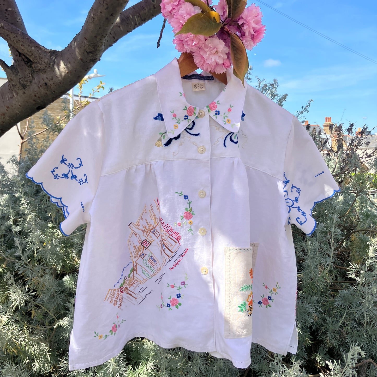 Short sleeved shirt made from a patchwork of reclaimed tablecloths with a traditional English pub with a thatched roof embroidered on the front and two bows on the yoke