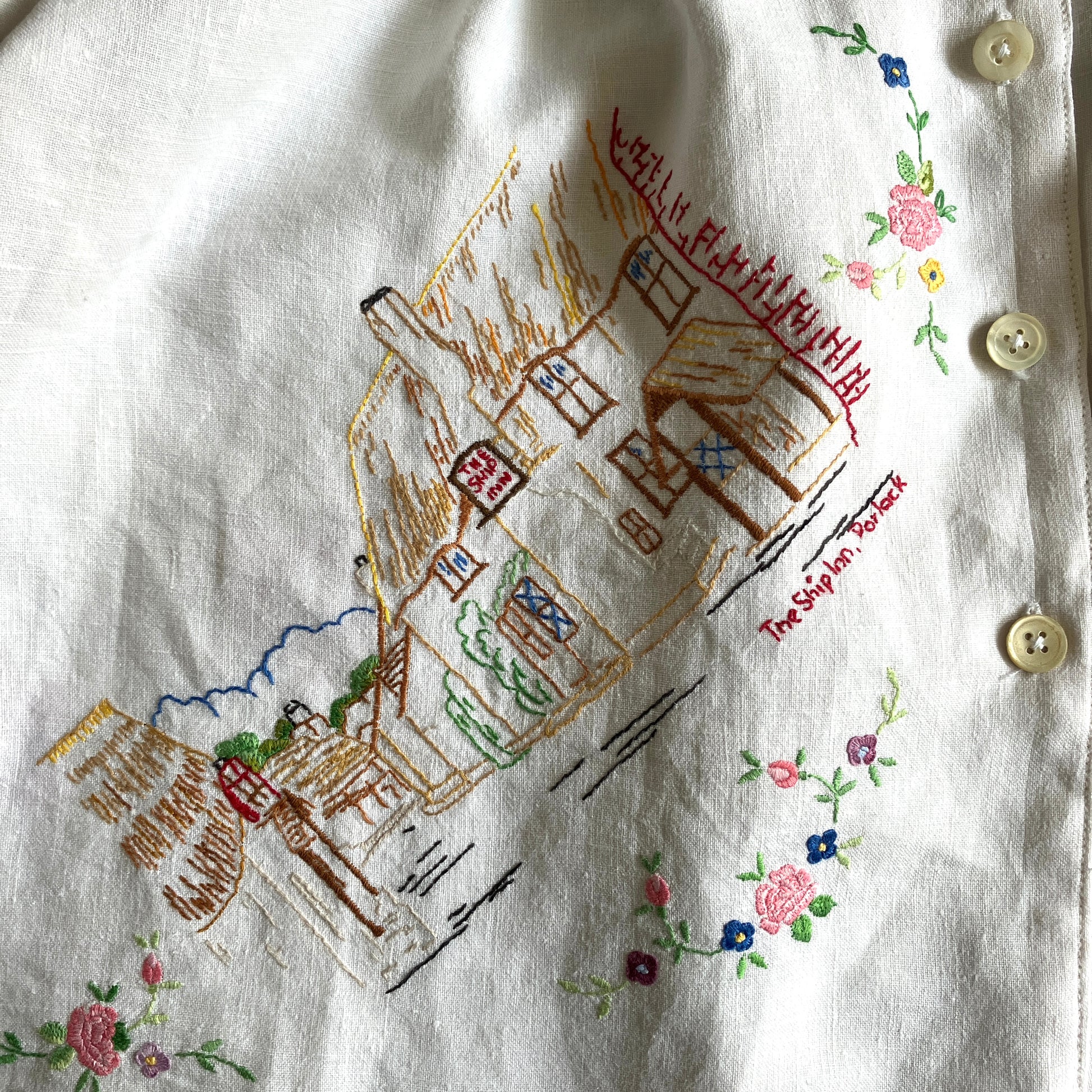 Short sleeved shirt made from a patchwork of reclaimed tablecloths with a traditional English pub with a thatched roof embroidered on the front and two bows on the yoke