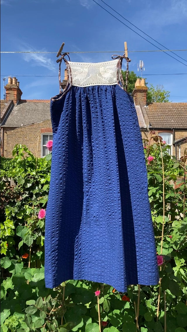 Hand stitched blue and white light and airy summer dress made from recycled cotton seersucker with a vintage bird-motif crochet yoke, tie straps and two patch pockets, hanging on a washing line, blowing in the breeze