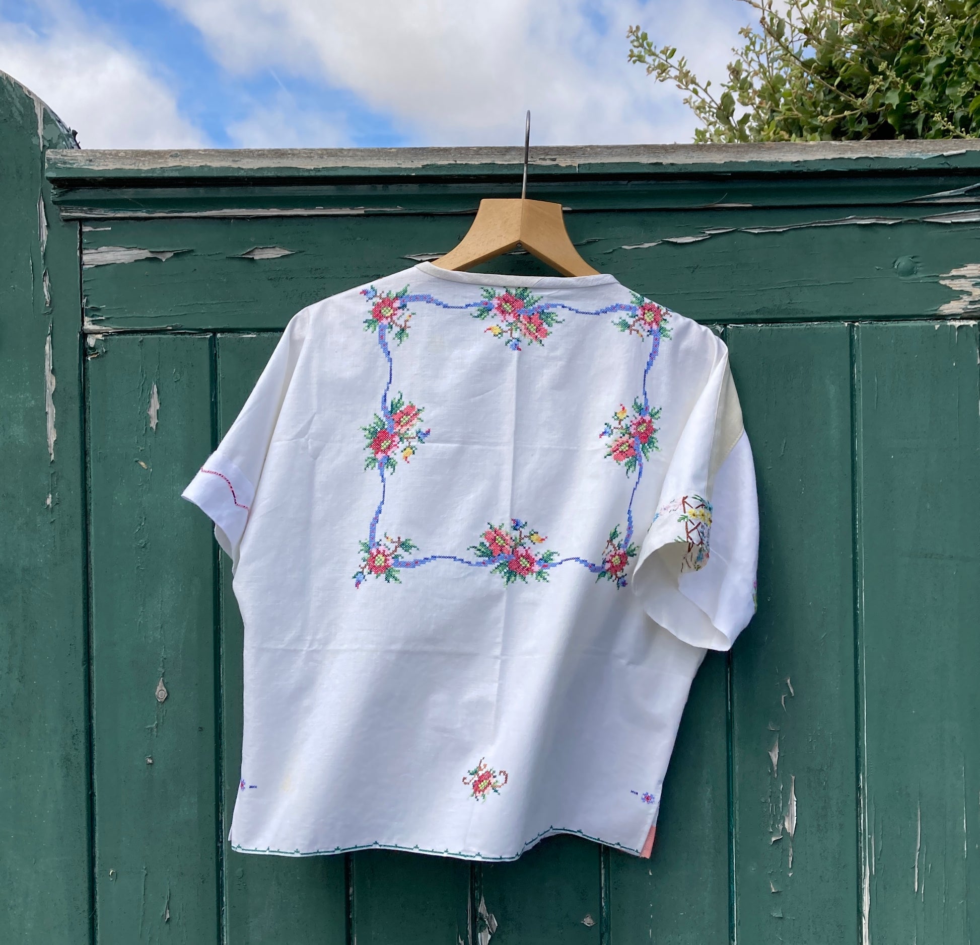Short-sleeved boxy top made from vintage embroidered linen tablecloths. Mostly white and off-white with wide pink stripes and embroidered details include cherries and flowers (back view)