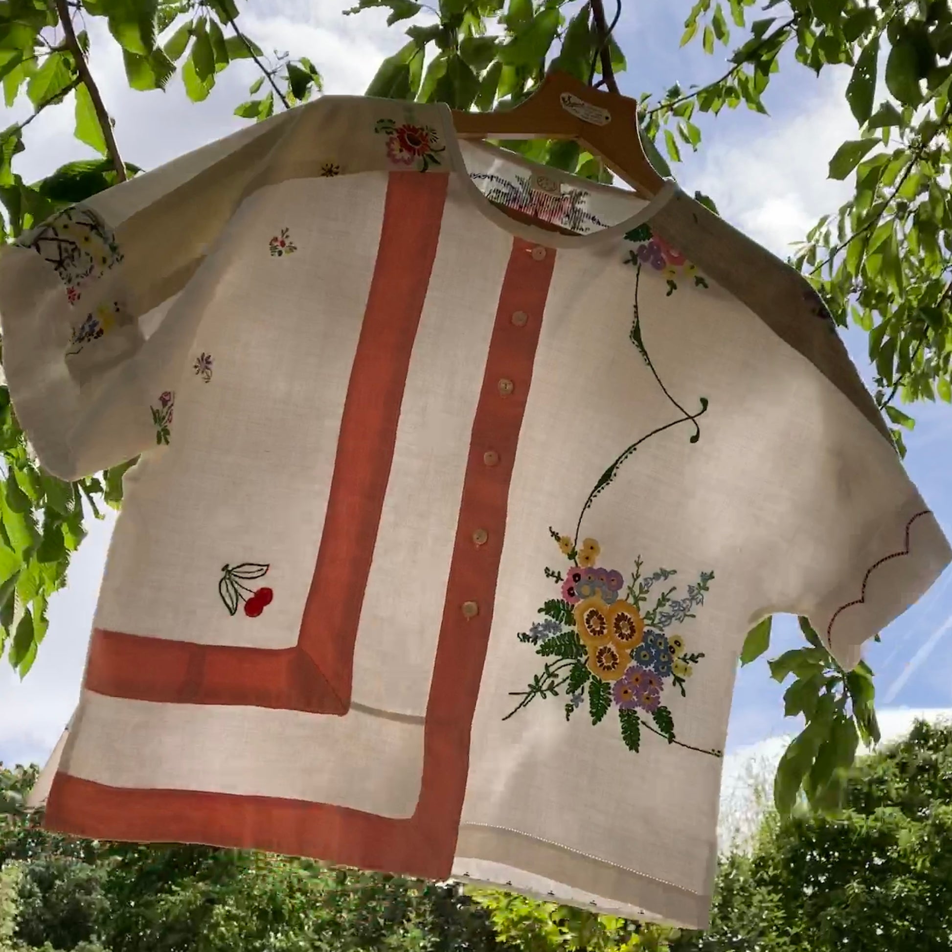 Short-sleeved boxy top made from vintage embroidered linen tablecloths. Mostly white and off-white with wide pink stripes and embroidered details include cherries and flowers. Pictured hanging in a tree