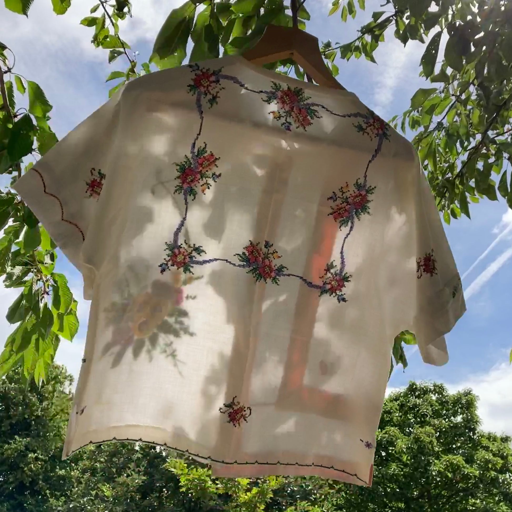 Short-sleeved boxy top made from vintage embroidered linen tablecloths. Mostly white and off-white with wide pink stripes and embroidered details include cherries and flowers. Pictured hanging in a tree (back view)