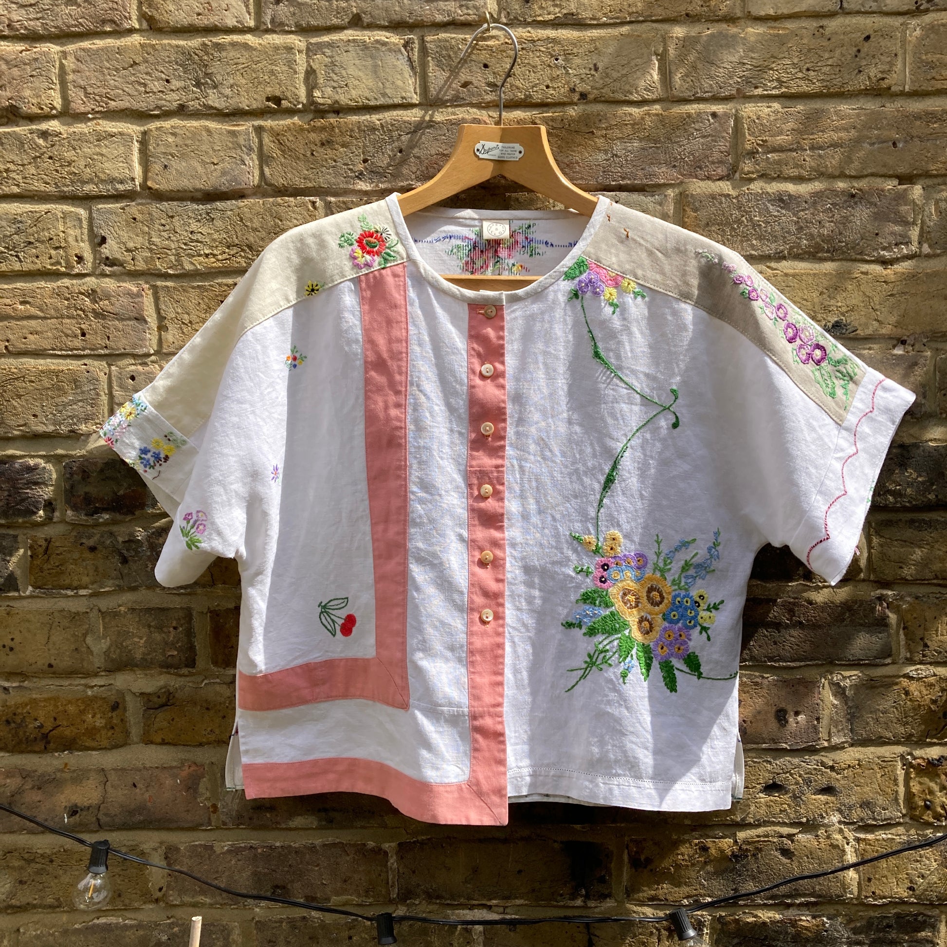 Short-sleeved boxy top made from vintage embroidered linen tablecloths. Mostly white and off-white with wide pink stripes and embroidered details include cherries and flowers
