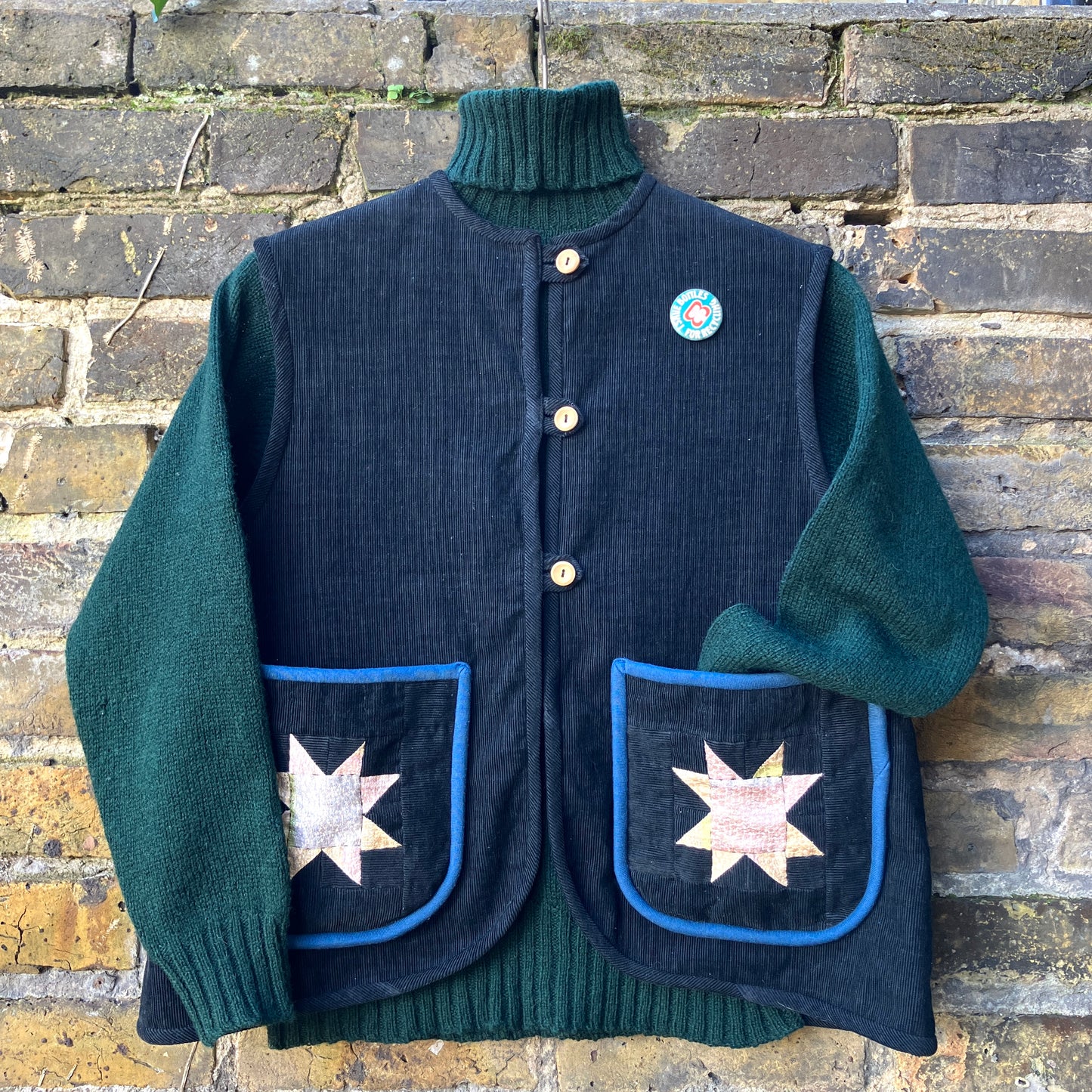 Needlecord vest/gilet/waistcoat with large hand quilted patchwork pockets in gold and silver lamé with blue binding. Finished with wooden buttons and a hanging loop. Pictured hanging against a brick wall with a dark green woollen jumper underneath