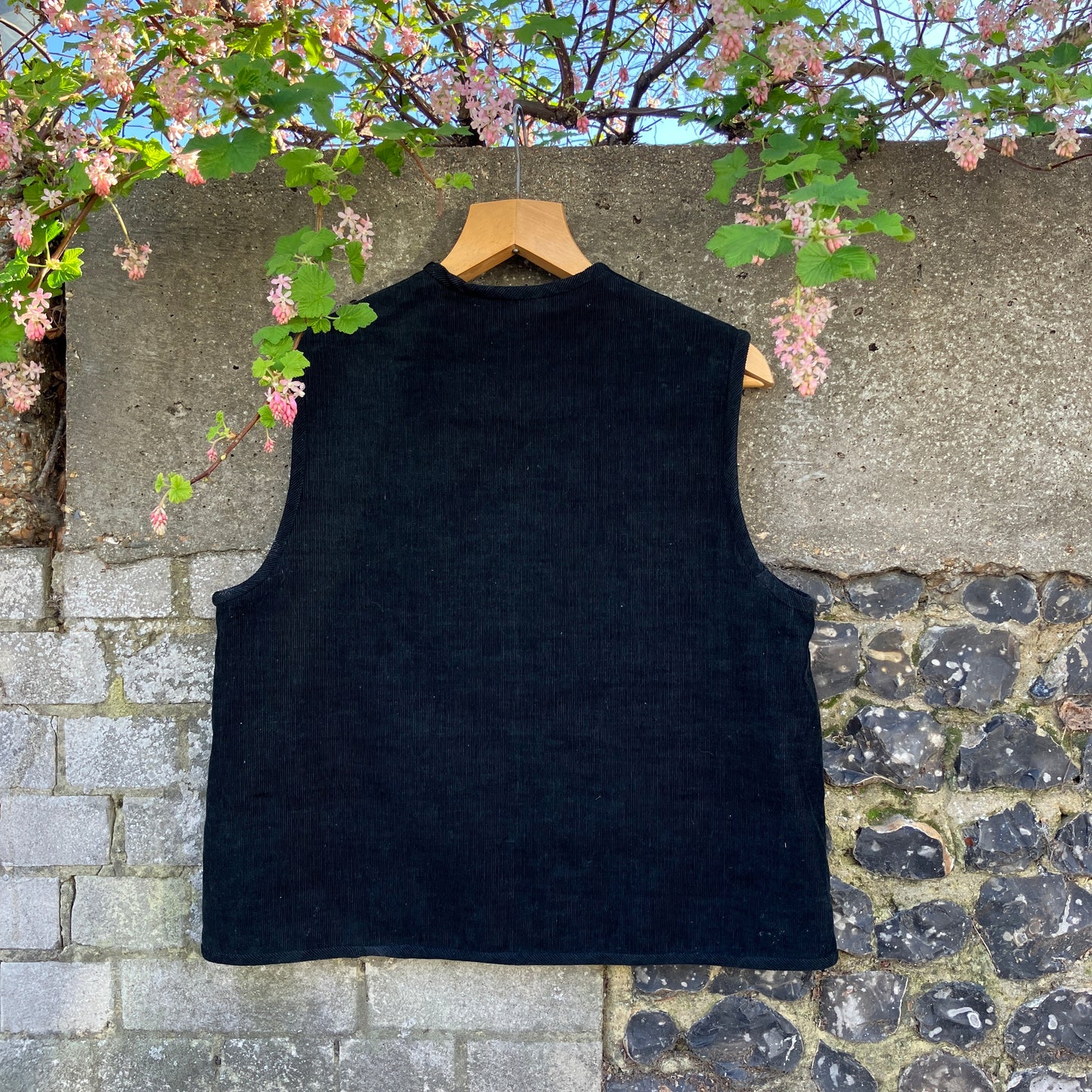 Needlecord vest/gilet/waistcoat with large hand quilted patchwork pockets in gold and silver lamé with blue binding. Finished with wooden buttons and a hanging loop. Pictured hanging against a wall with spring blossom overhanging (back view)