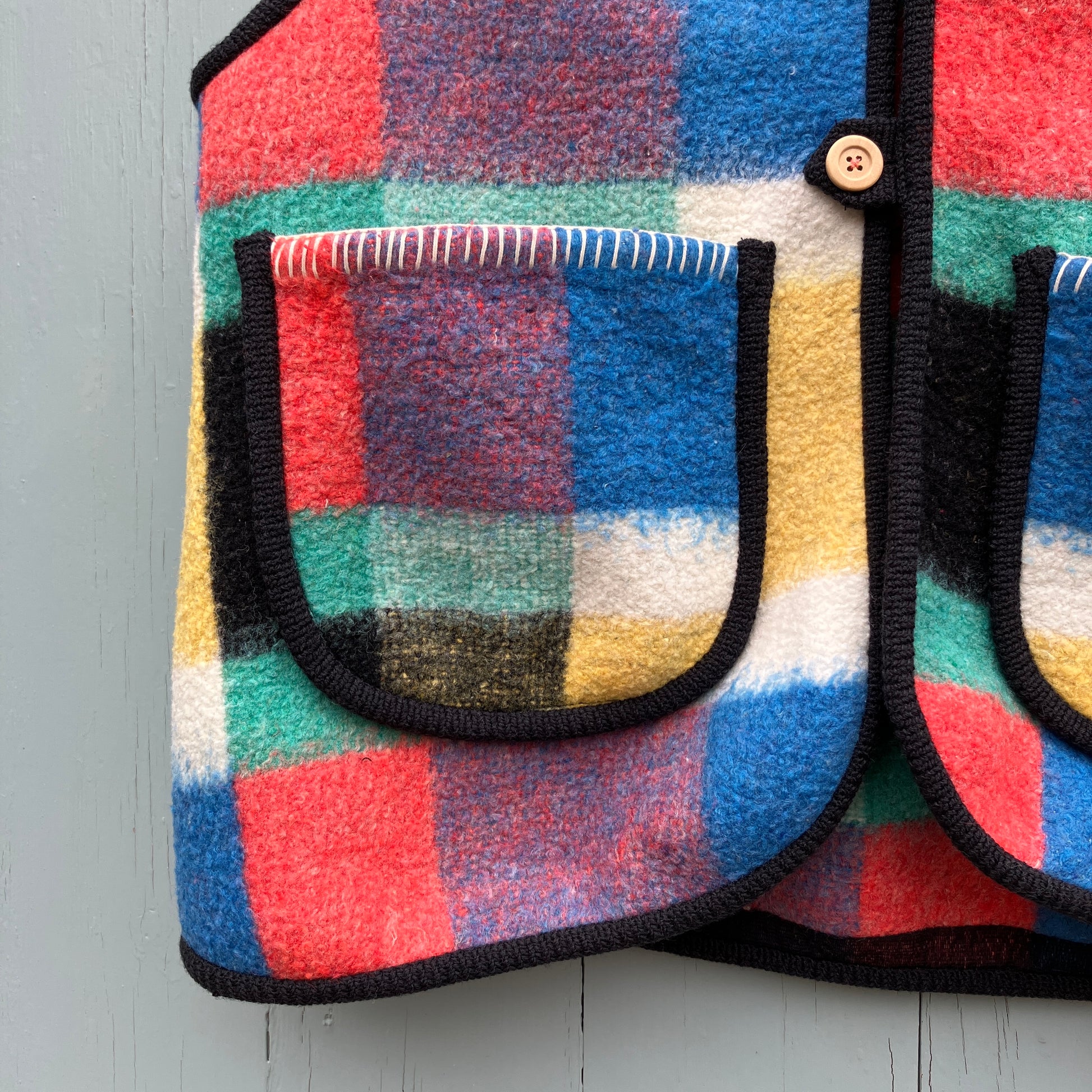 A vest/gilet/waistcoat made from a multicoloured checked reclaimed blanket. It fastens with three wooden buttons and has to large patch pockets (close-up)