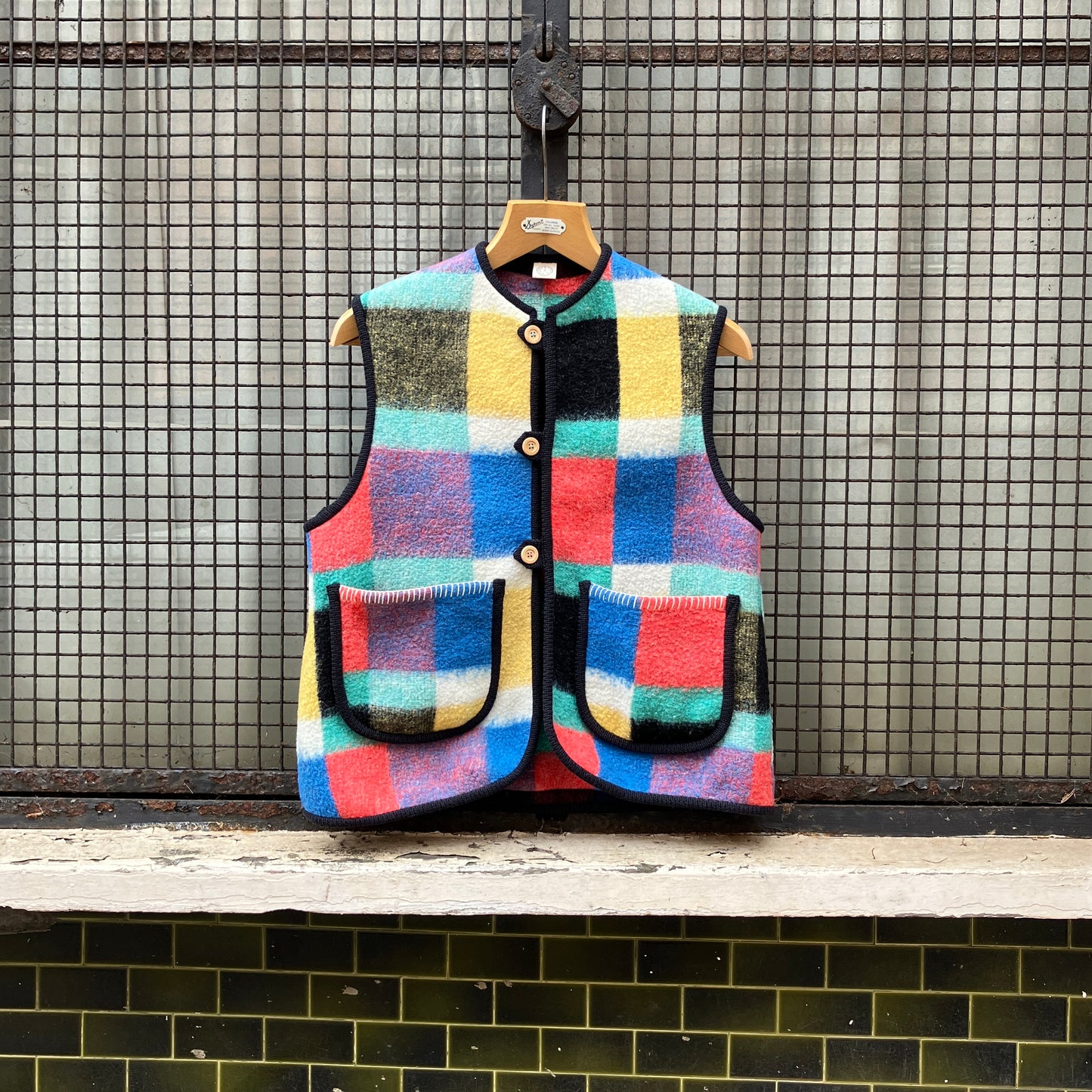 A vest/gilet/waistcoat made from a multicoloured checked reclaimed blanket. It fastens with three wooden buttons and has to large patch pockets