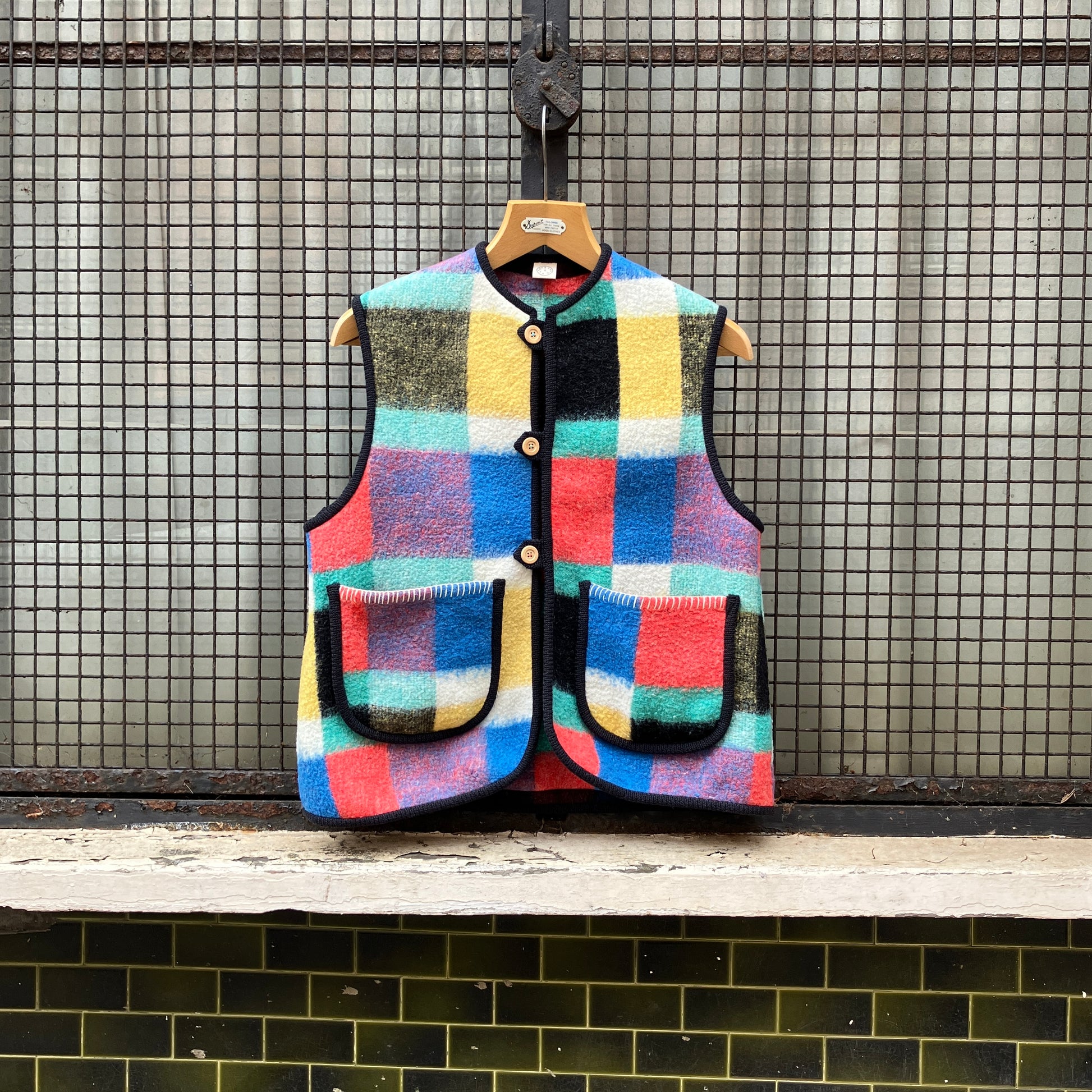 A vest/gilet/waistcoat made from a multicoloured checked reclaimed blanket. It fastens with three wooden buttons and has to large patch pockets