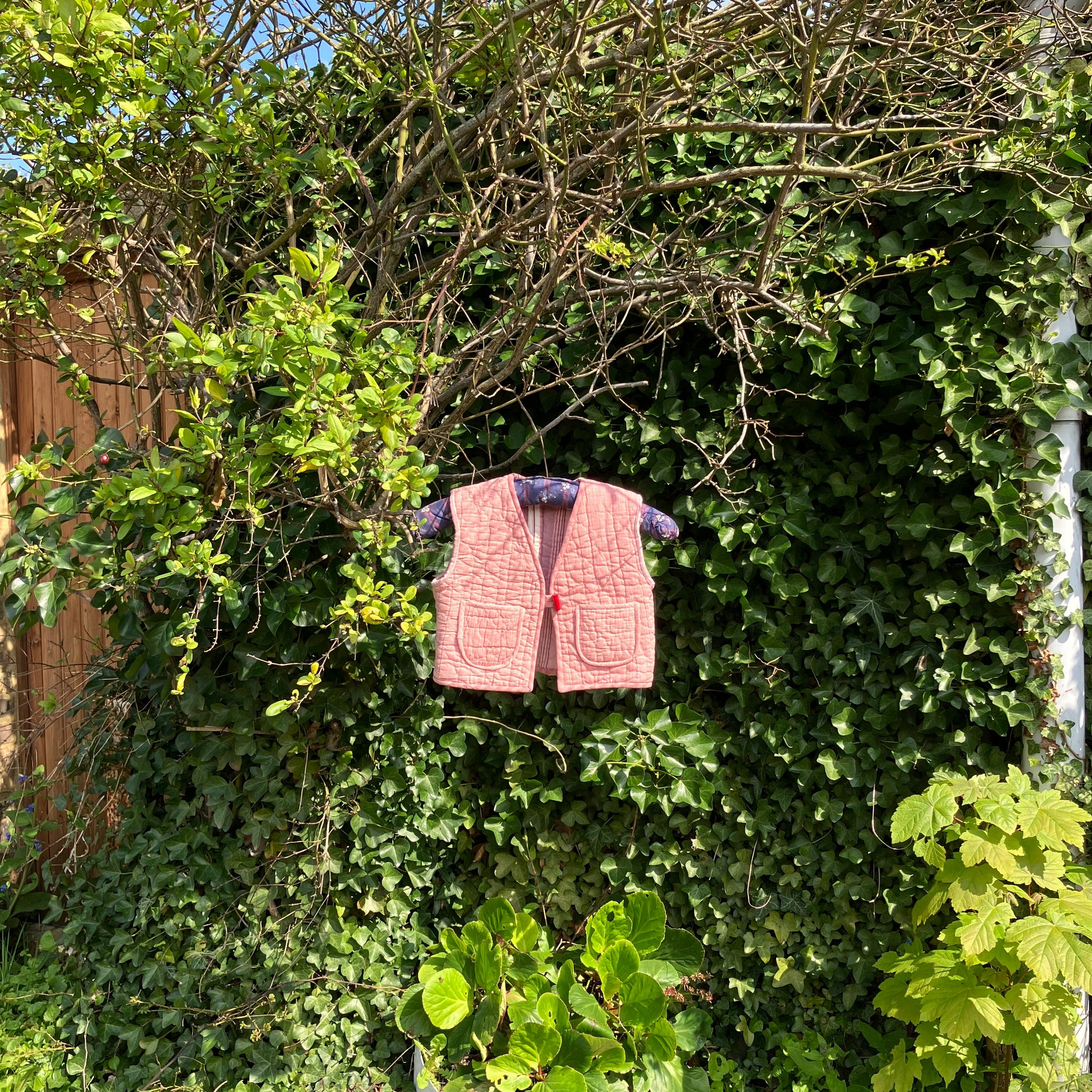 kids vest/gilet/waistcoat made from offcuts of a reclaimed pink quilt. Shown hanging on a blue vintage hanger in some foliage