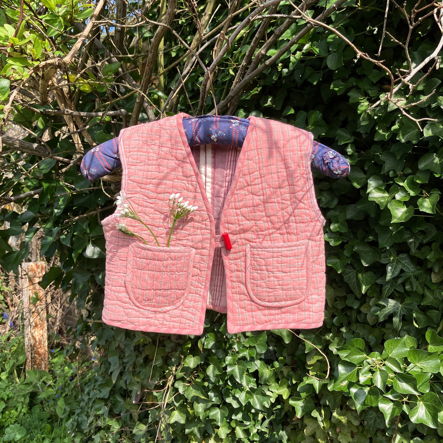 kids vest/gilet/waistcoat made from offcuts of a reclaimed pink quilt. Shown hanging on a blue vintage hanger in some foliage with some wild flowers in one of the pockets