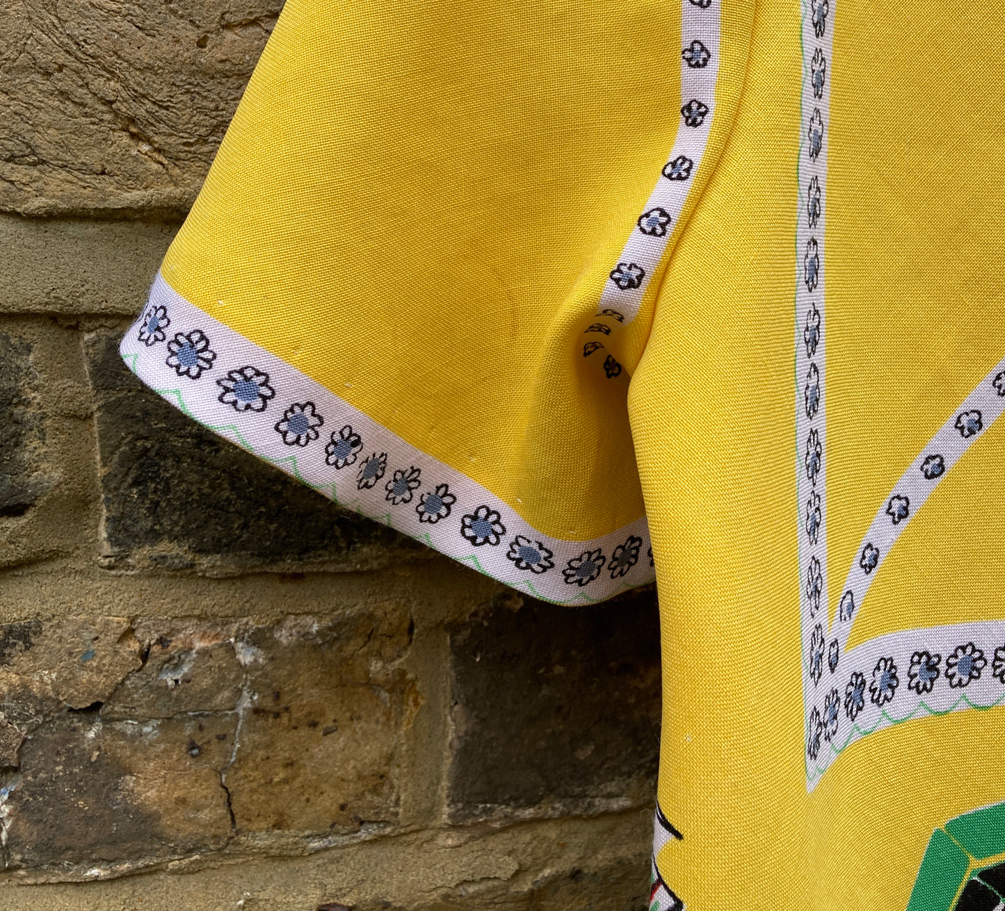 short-sleeved shirt made from a recycled tablecloth printed with picnic food a record player and records, mainly yellow and white, hanging on a hanger against a brick wall (mark on sleeve)