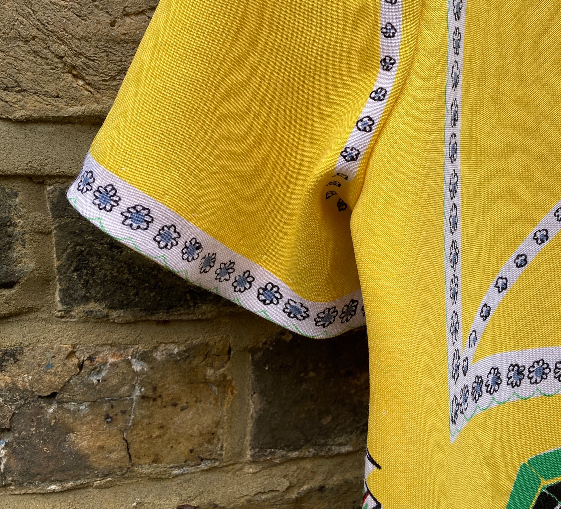 short-sleeved shirt made from a recycled tablecloth printed with picnic food a record player and records, mainly yellow and white, hanging on a hanger against a brick wall (mark on sleeve)