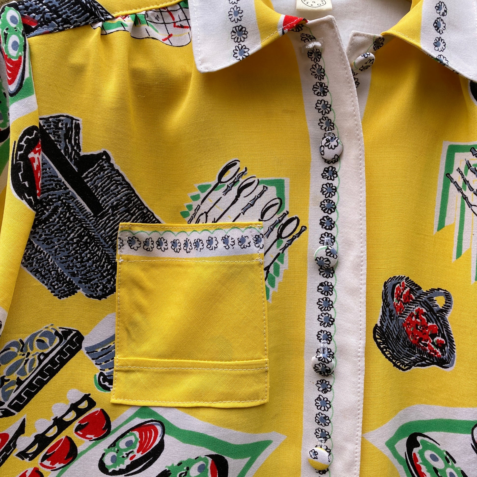 short-sleeved shirt made from a recycled tablecloth printed with picnic food a record player and records, mainly yellow and white, hanging on a hanger against a brick wall (small marks on front)