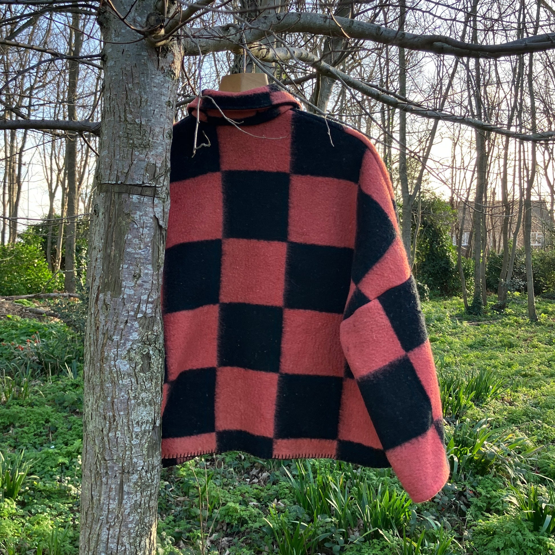 Back view of a pink and black checkerboard blanket jacket handmade from a reclaimed blanket. Pictured hanging in a tree