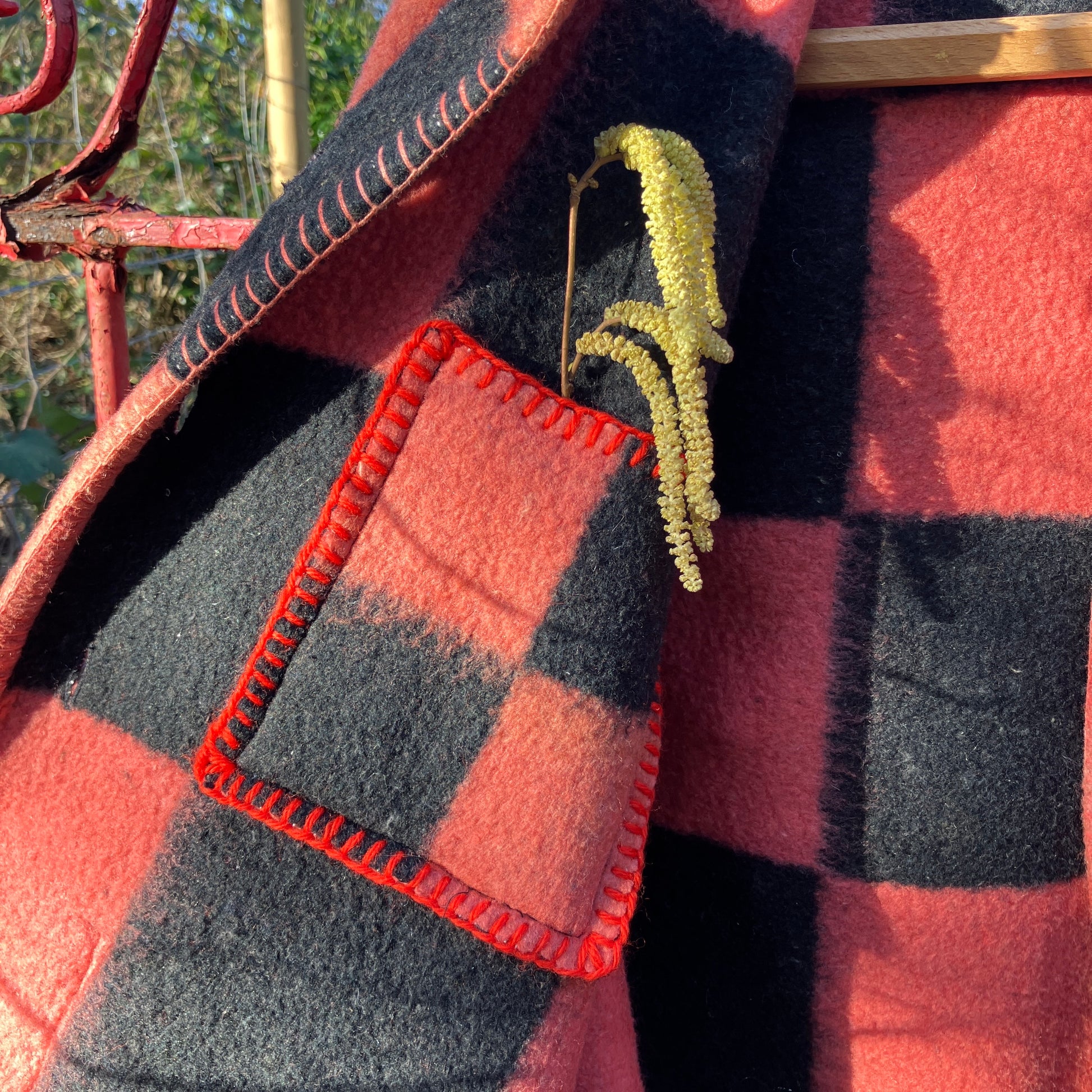 Close-up of the inside pocket of a pink and black checkerboard blanket jacket handmade from a reclaimed blanket. The pocket is finished with red blanket stitch and there is a catkin in