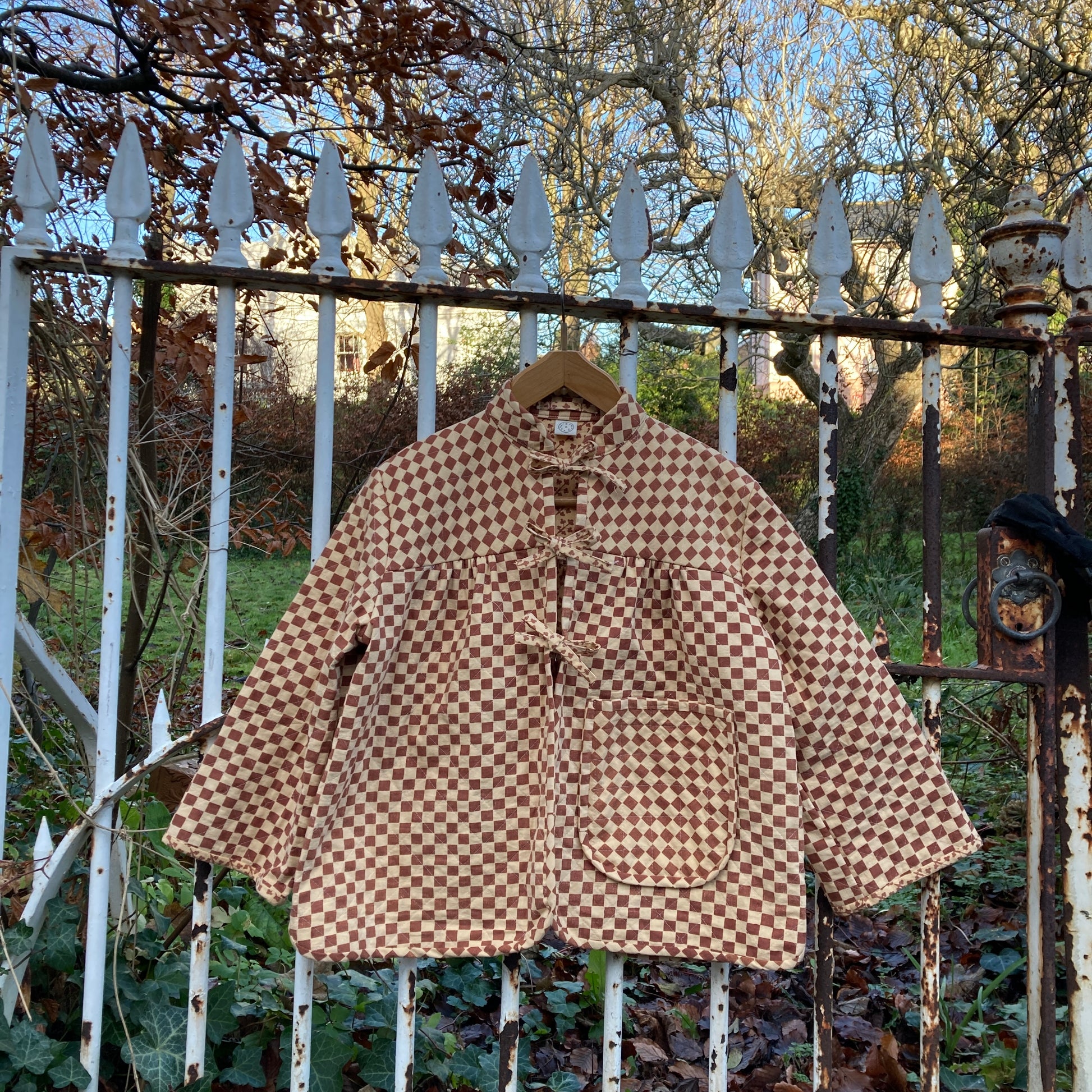 A quilted jacket in two shades of brown checkerboard print that fastens with three bows down the front. It has a big pocket and its hanging on an ornate but rusty gate