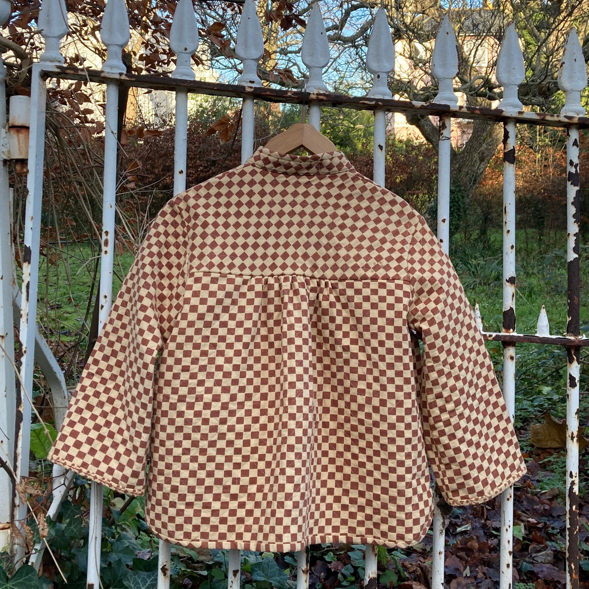 A quilted jacket in two shades of brown checkerboard print that fastens with three bows down the front. It has a big pocket and its hanging on an ornate but rusty gate (back view)