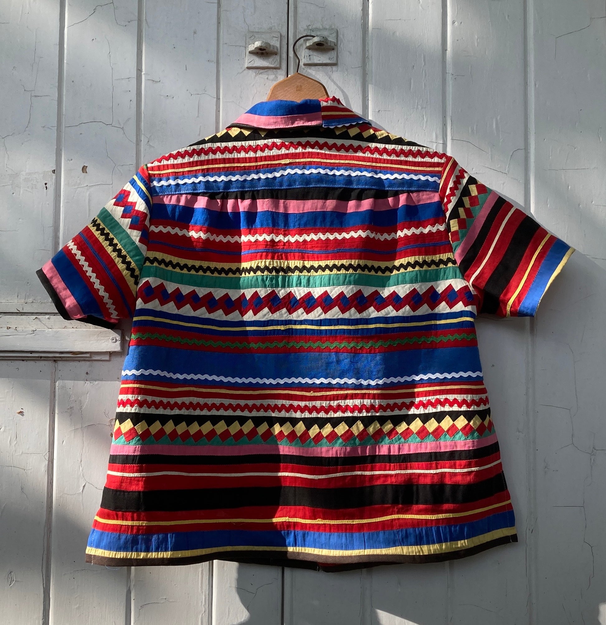 A short-sleeved shirt made from brightly coloured seminole patchwork hanging on a hanger on a white painted door (back view)