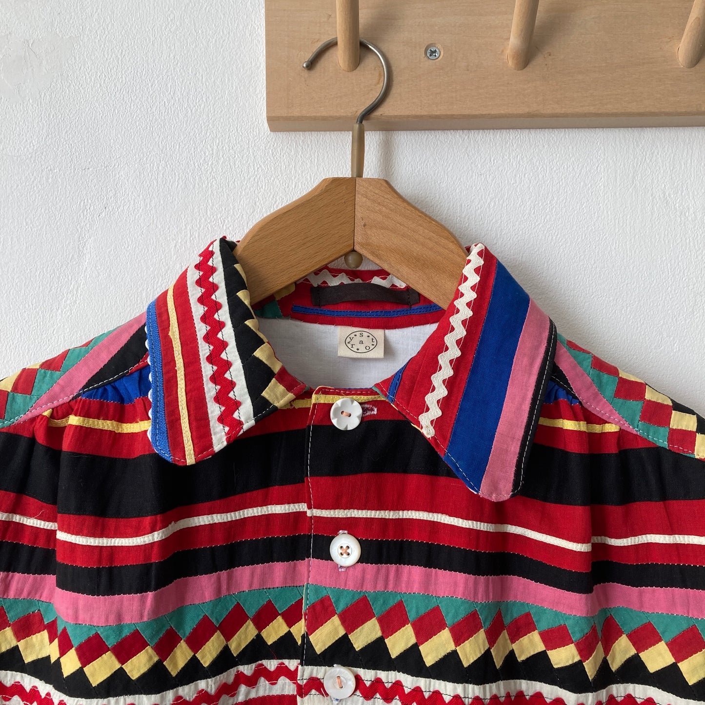 A short-sleeved shirt made from brightly coloured seminole patchwork hanging on a hanger from some wooden hooks (close up)