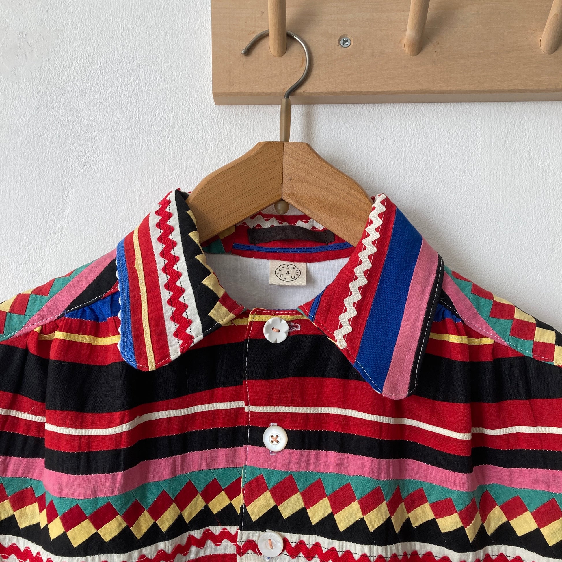 A short-sleeved shirt made from brightly coloured seminole patchwork hanging on a hanger from some wooden hooks (close up)