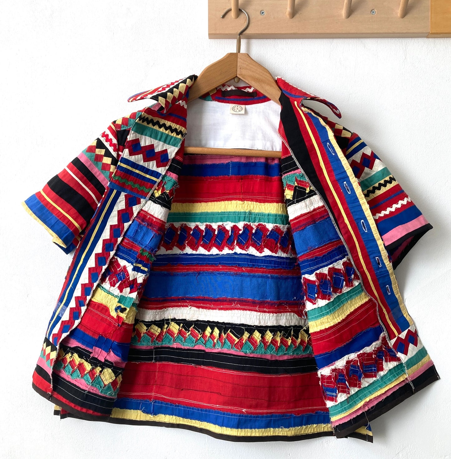 A short-sleeved shirt made from brightly coloured seminole patchwork hanging on a hanger from some wooden hooks (shows inside)