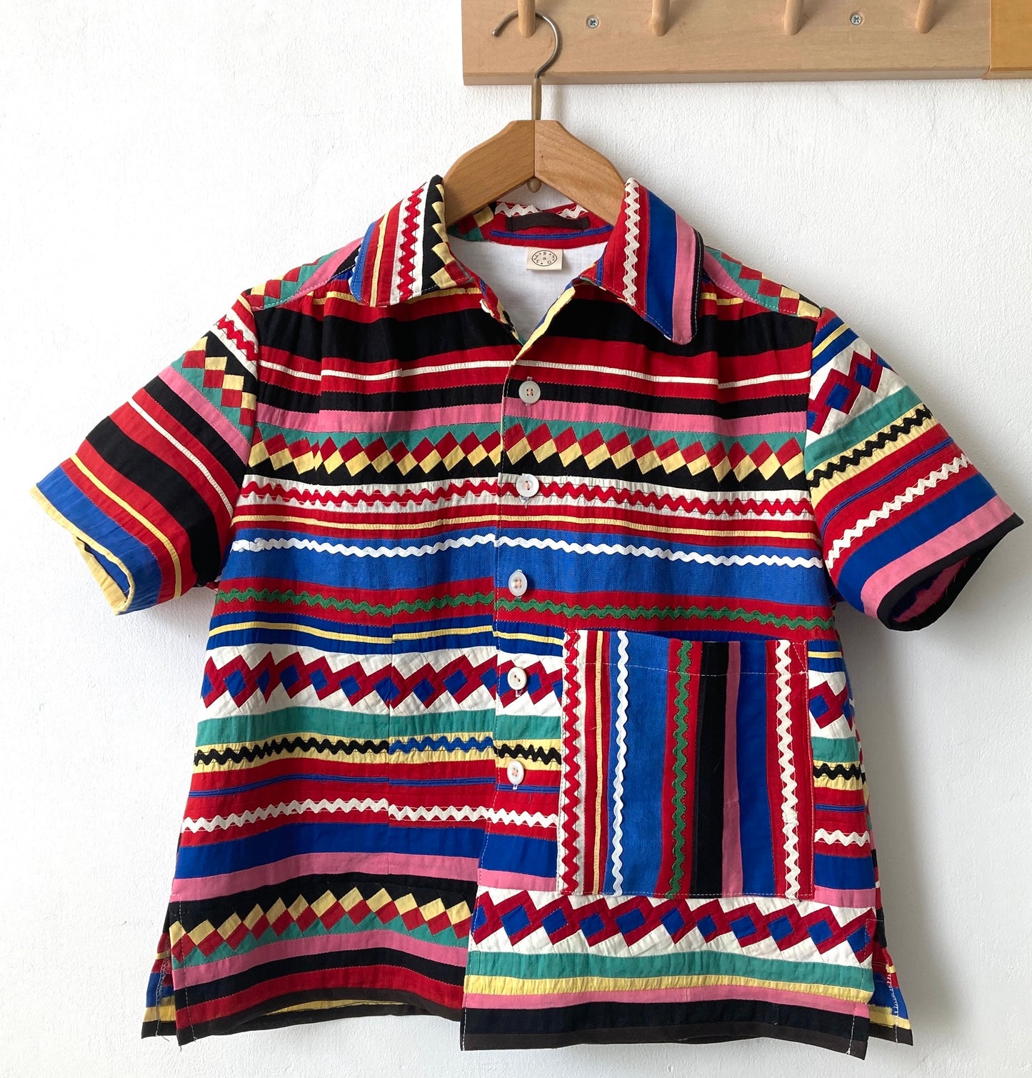 A short-sleeved shirt made from brightly coloured seminole patchwork hanging on a hanger from some wooden hooks