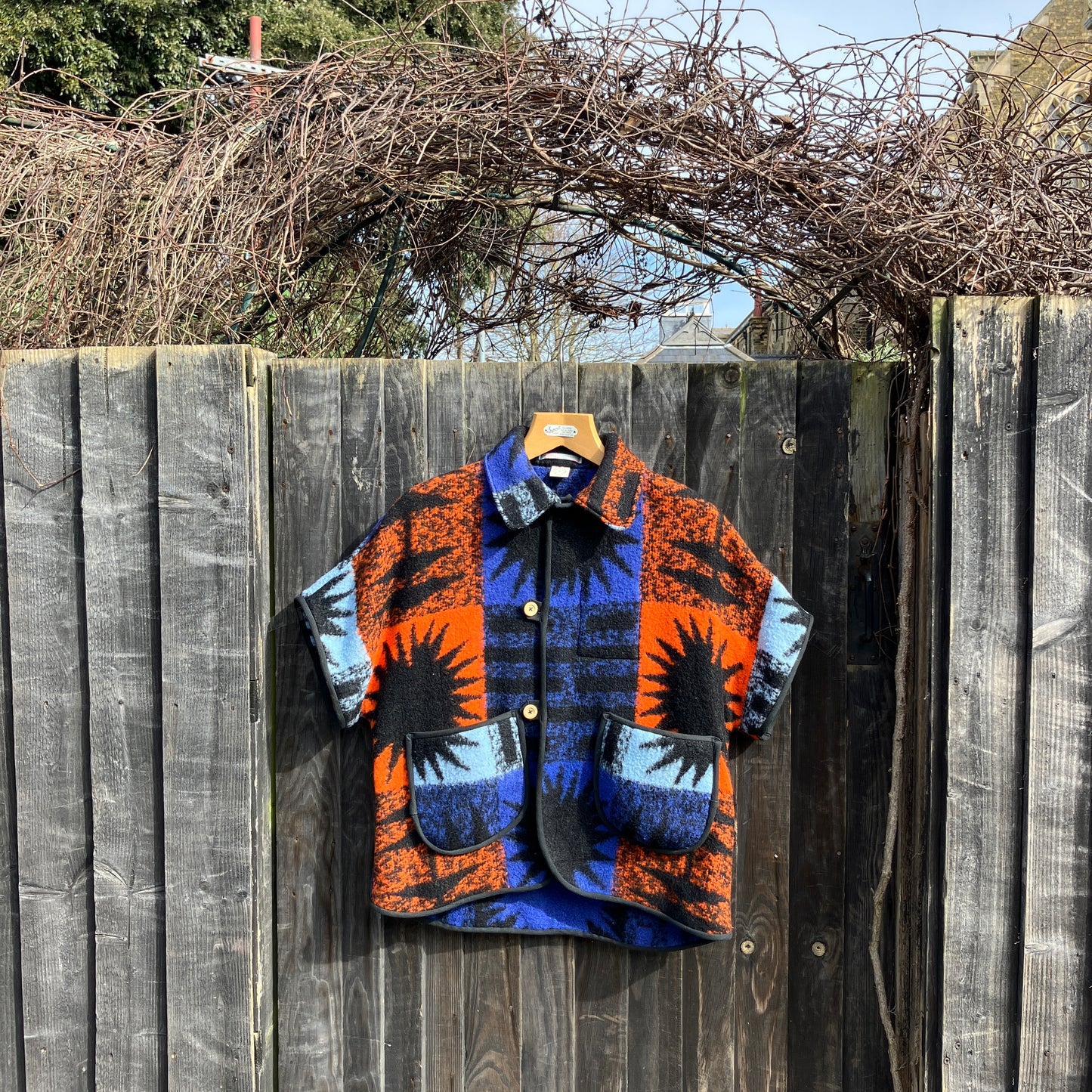 Crop-sleeved jacket made from a fabulous reclaimed blanket with starburst motifs in bright blue, light blue, black and orange, hanging on a garden fence