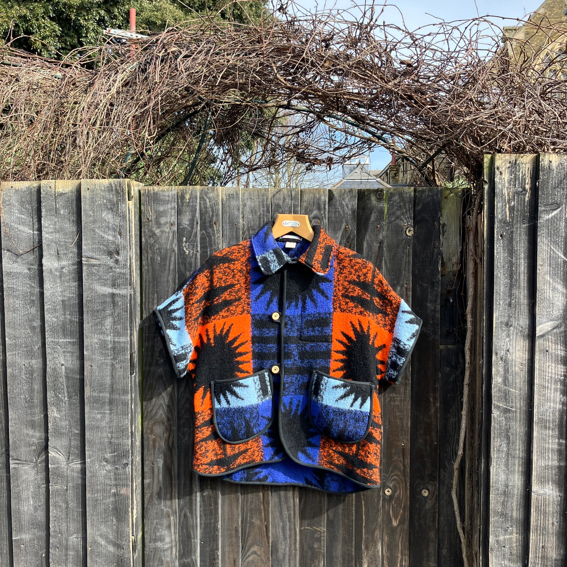 Crop-sleeved jacket made from a fabulous reclaimed blanket with starburst motifs in bright blue, light blue, black and orange, hanging on a garden fence
