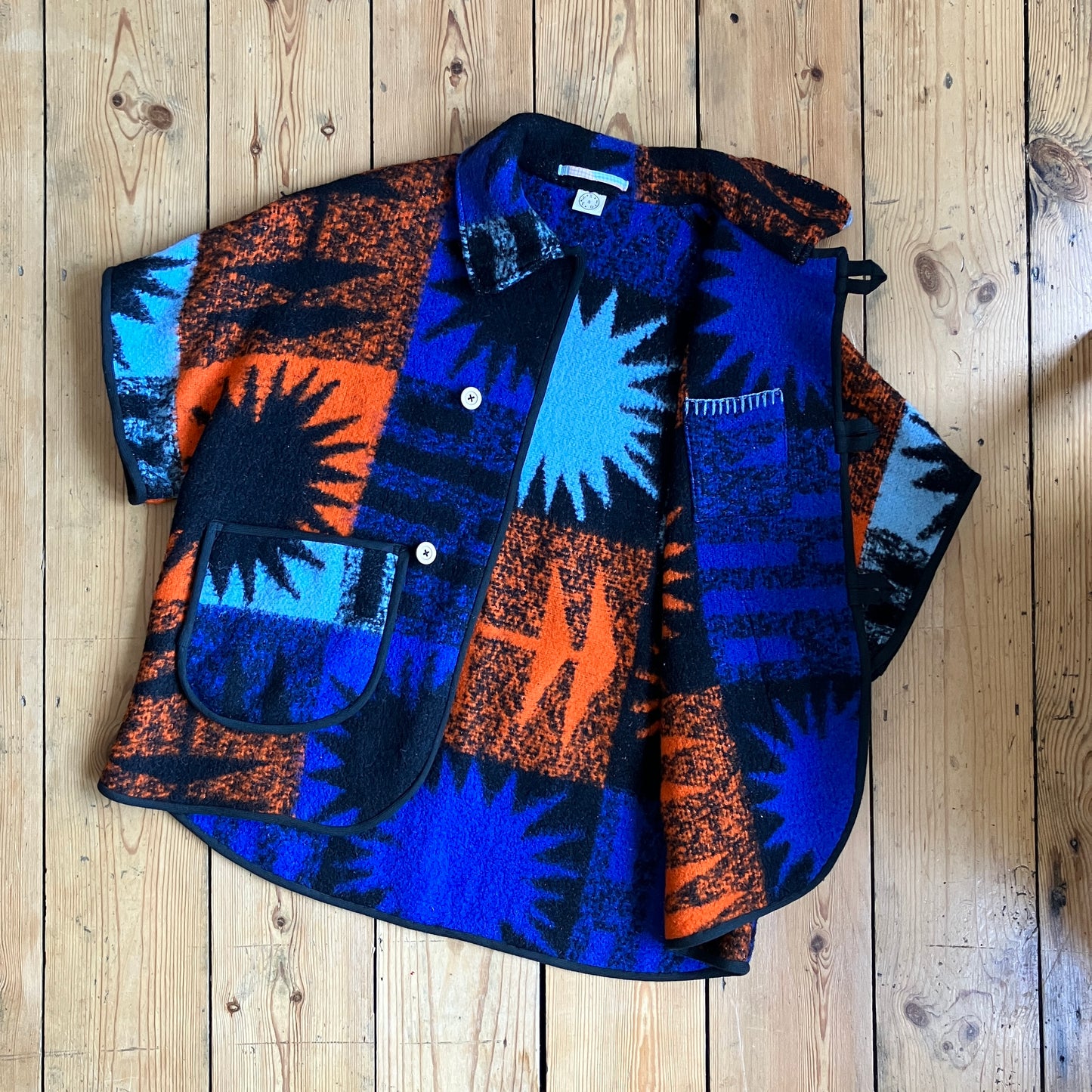 Crop-sleeved jacket made from a fabulous reclaimed blanket with starburst motifs in bright blue, light blue, black and orange, shown on a wooden floor with the inside, which has a little hidden pocket visible