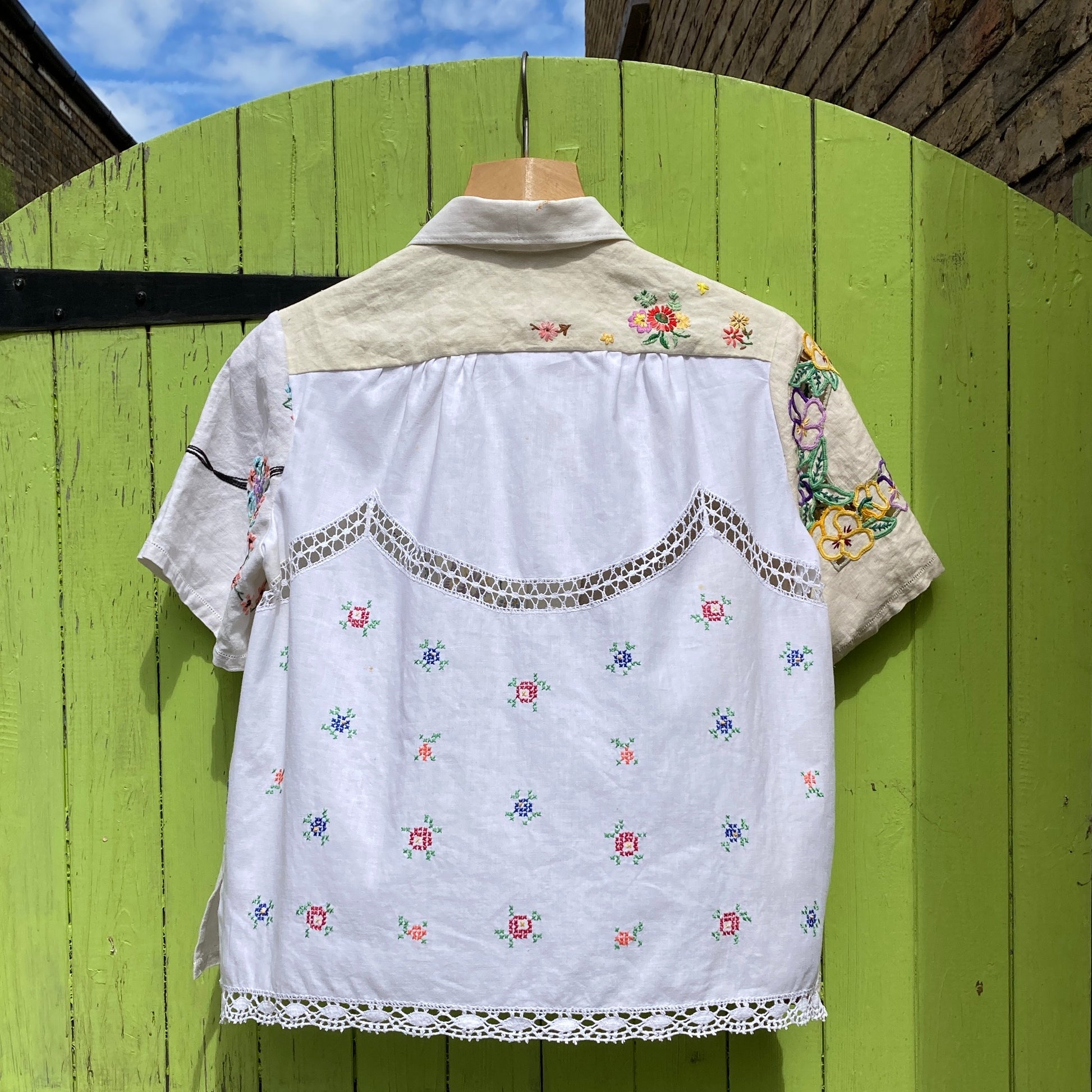 Short-sleeved shirt made from vintage hand embroidered tablecloths, mainly white and off-white with colourful embroidery. Photographed hanging on a green gate (back view)
