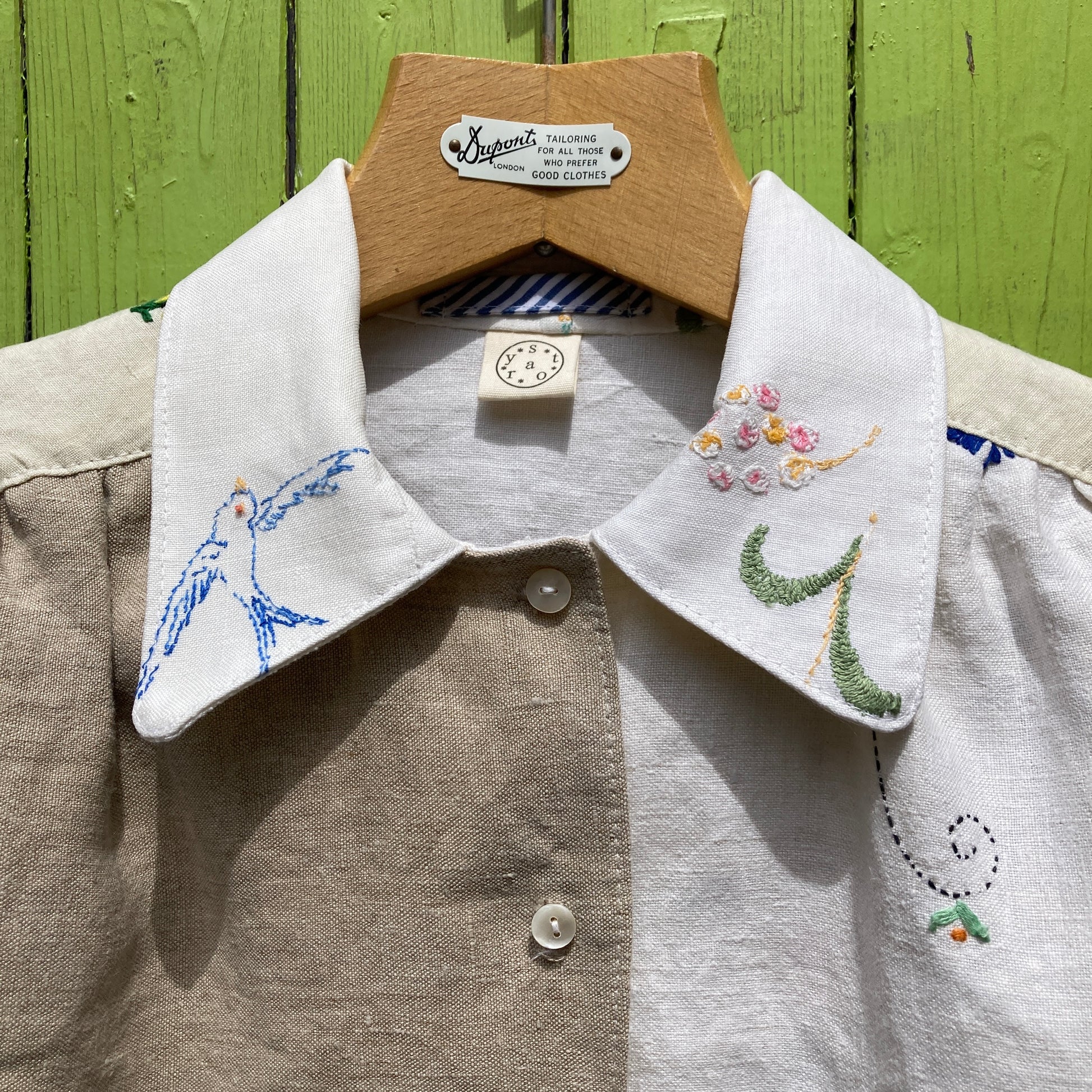 Short-sleeved shirt made from vintage hand embroidered tablecloths, mainly white and off-white with colourful embroidery. Photographed hanging on a green gate (close-up)