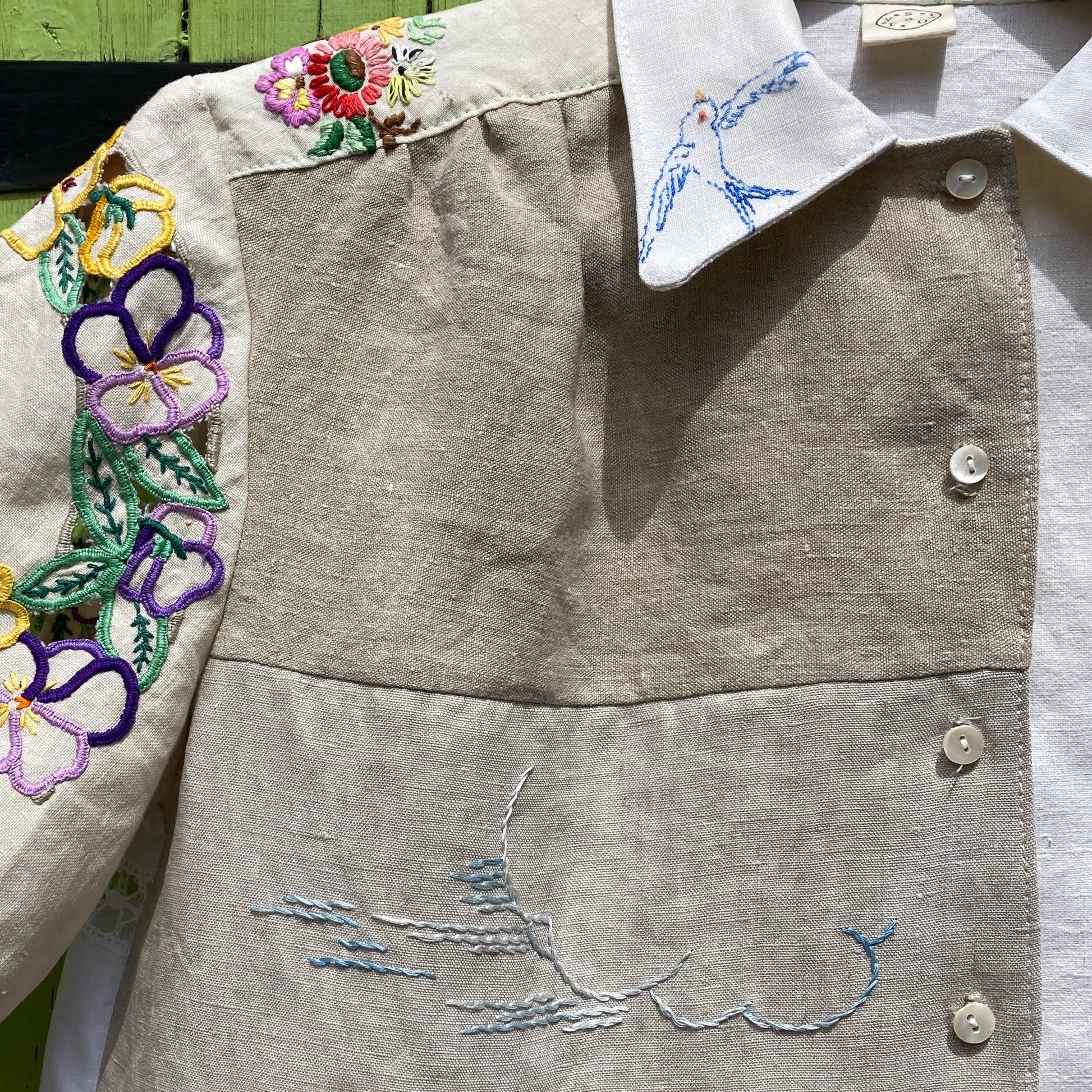 Short-sleeved shirt made from vintage hand embroidered tablecloths, mainly white and off-white with colourful embroidery. Photographed hanging on a green gate (close-up of cloud embroidery)