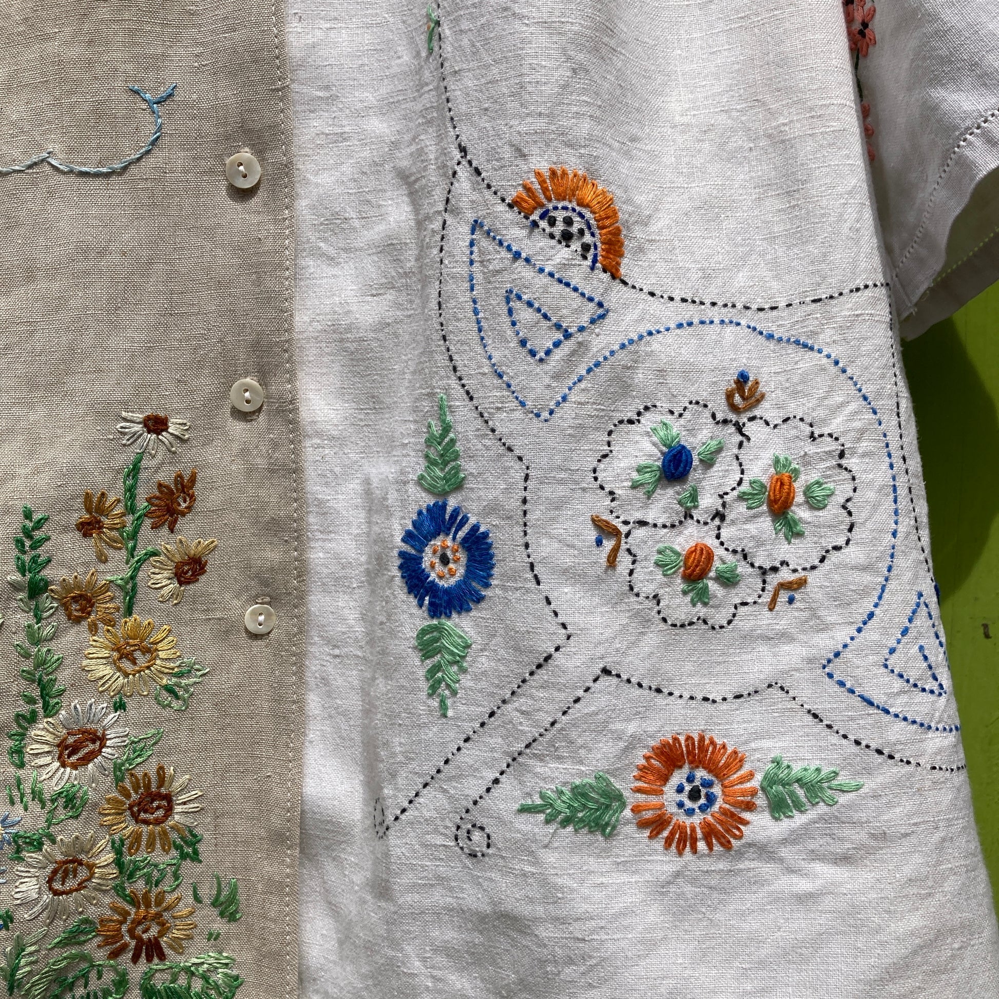 Short-sleeved shirt made from vintage hand embroidered tablecloths, mainly white and off-white with colourful embroidery. Photographed hanging on a green gate (close-up of floral embroidery)