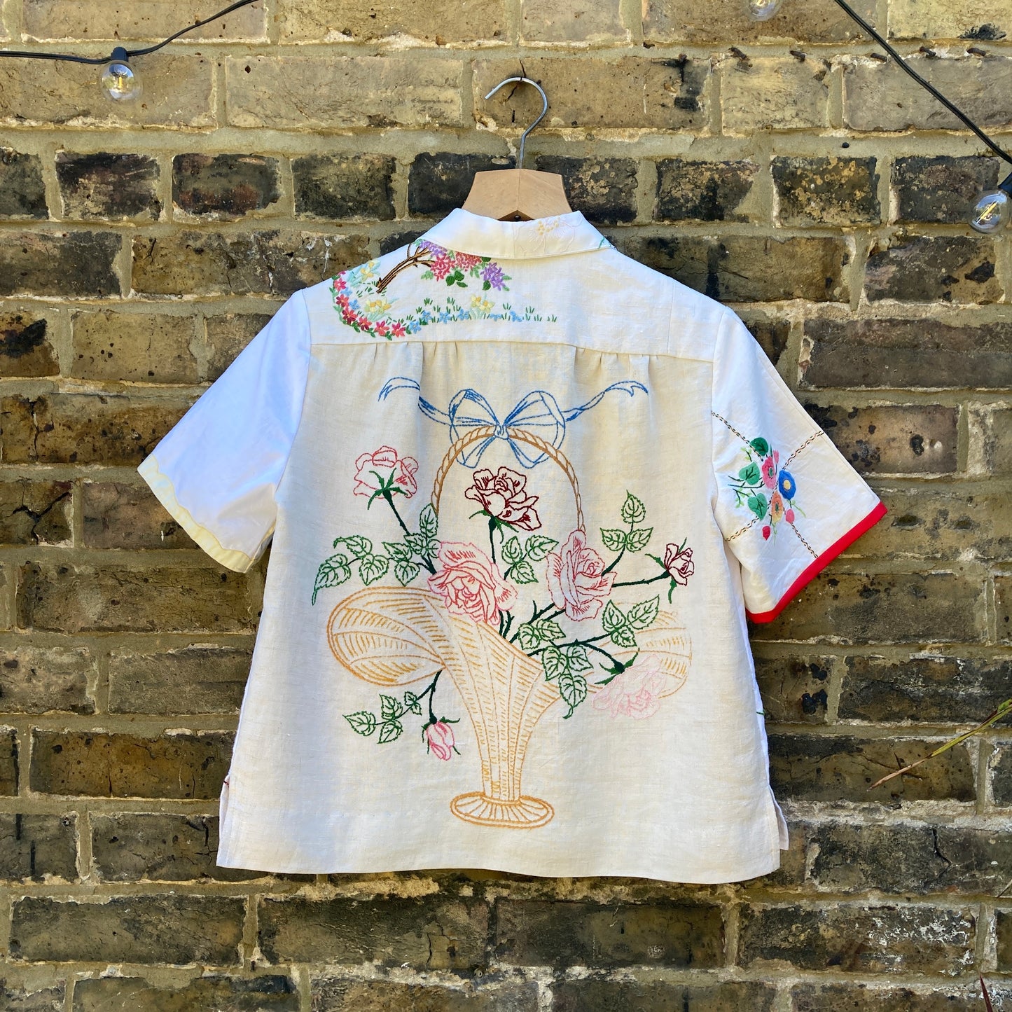 Back view of a short-sleeved shirt made from recycled tablecloths is mainly white and off-white with colourful embroidered and appliquéd flowers and orange buttons