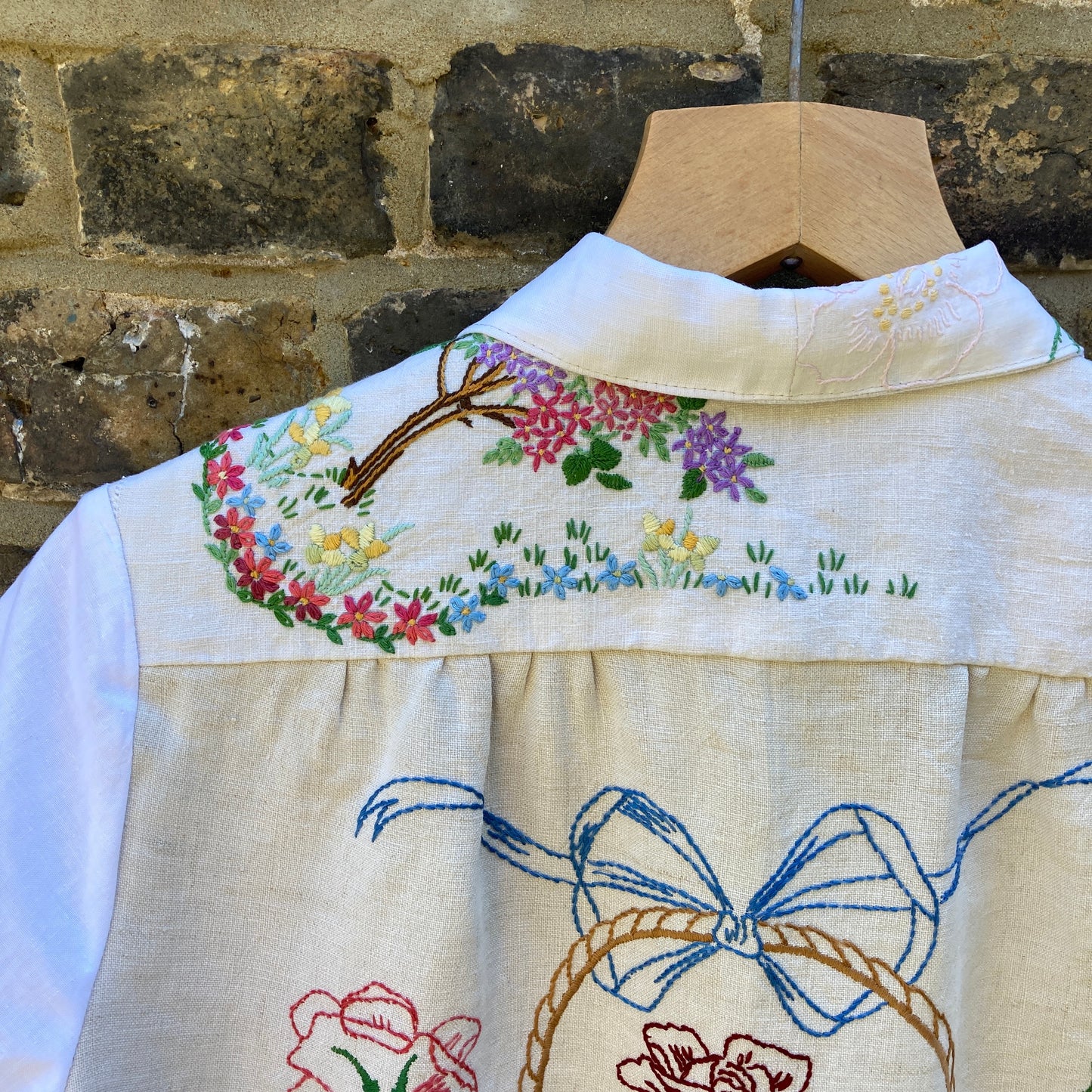 Close-up of back of short-sleeved shirt made from recycled tablecloths is mainly white and off-white with colourful embroidered and appliquéd flowers and orange buttons