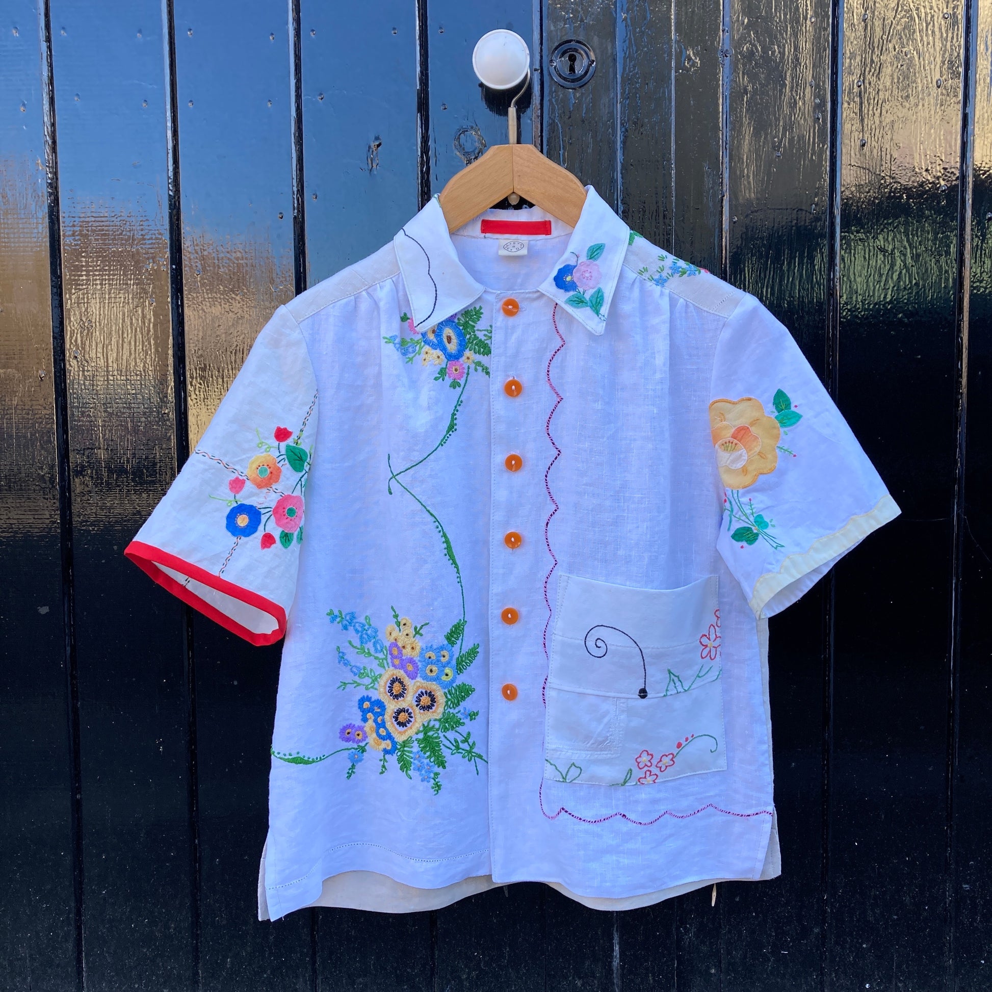 short-sleeved shirt made from recycled tablecloths is mainly white and off-white with colourful embroidered and appliquéd flowers and orange buttons